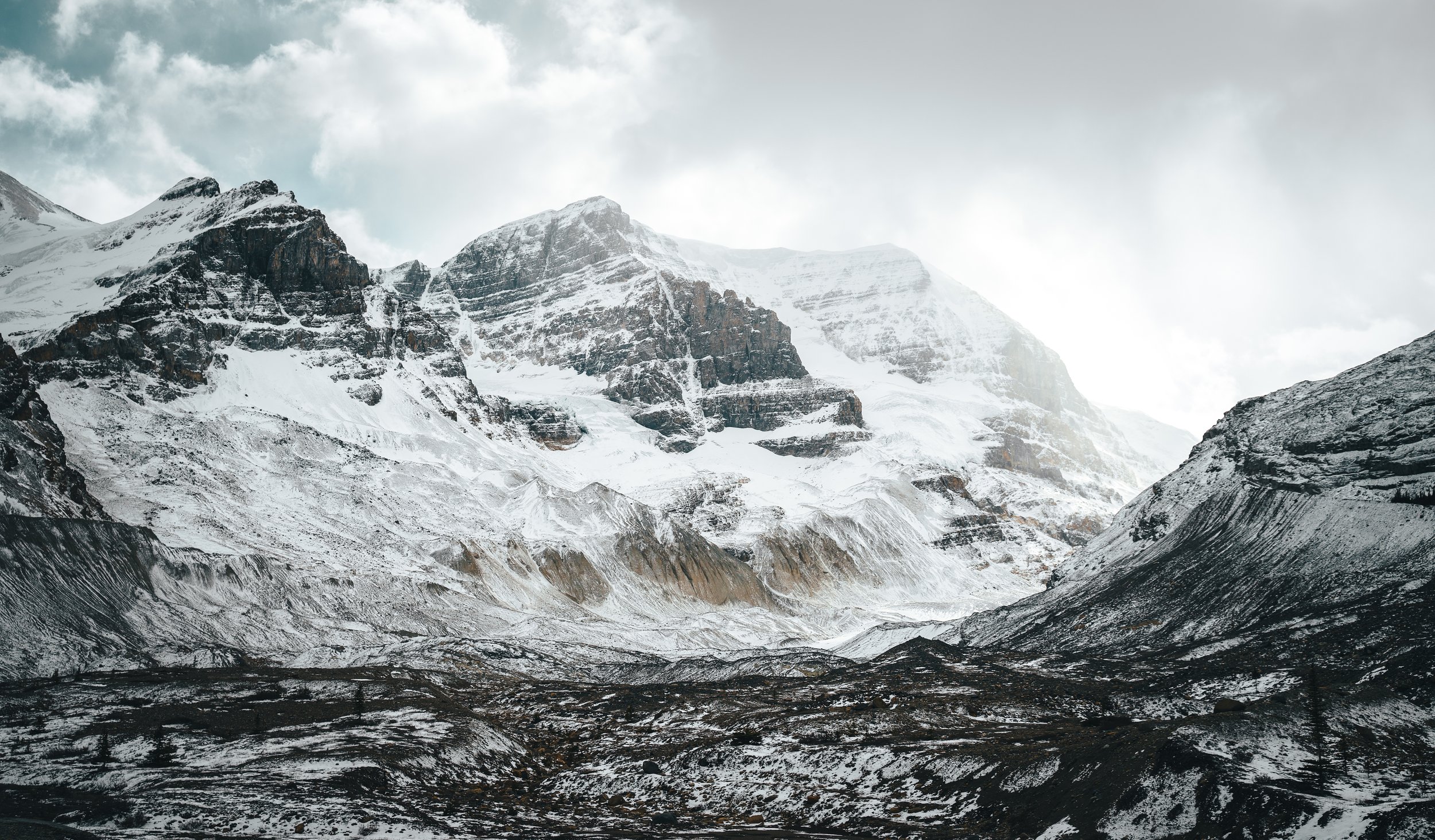 Snow-capped mountains under cloudy sky with rugged terrain in the foreground.