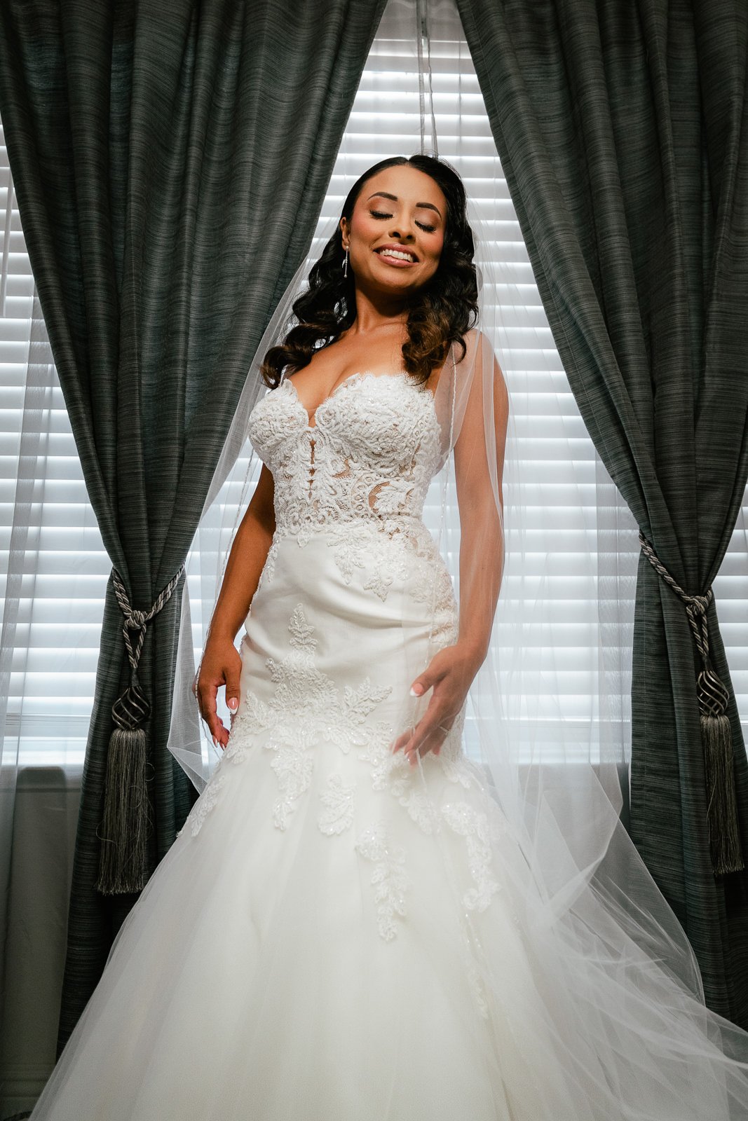 Bride smiling in lace wedding dress with veil, standing in front of curtains.