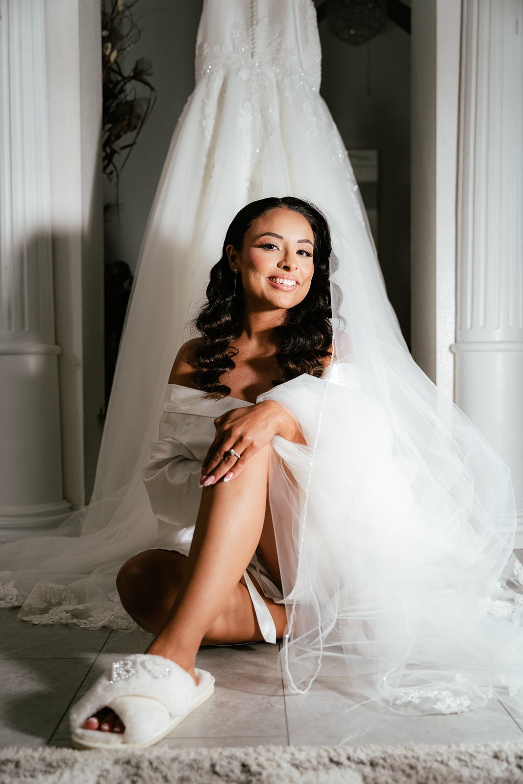 Bride sitting on the floor, wearing a robe and slippers, smiling with a wedding dress hanging in the background.