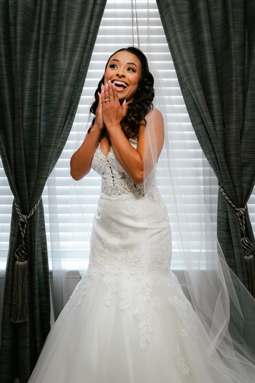 Bride in white lace wedding dress with veil, smiling in front of window with curtains.