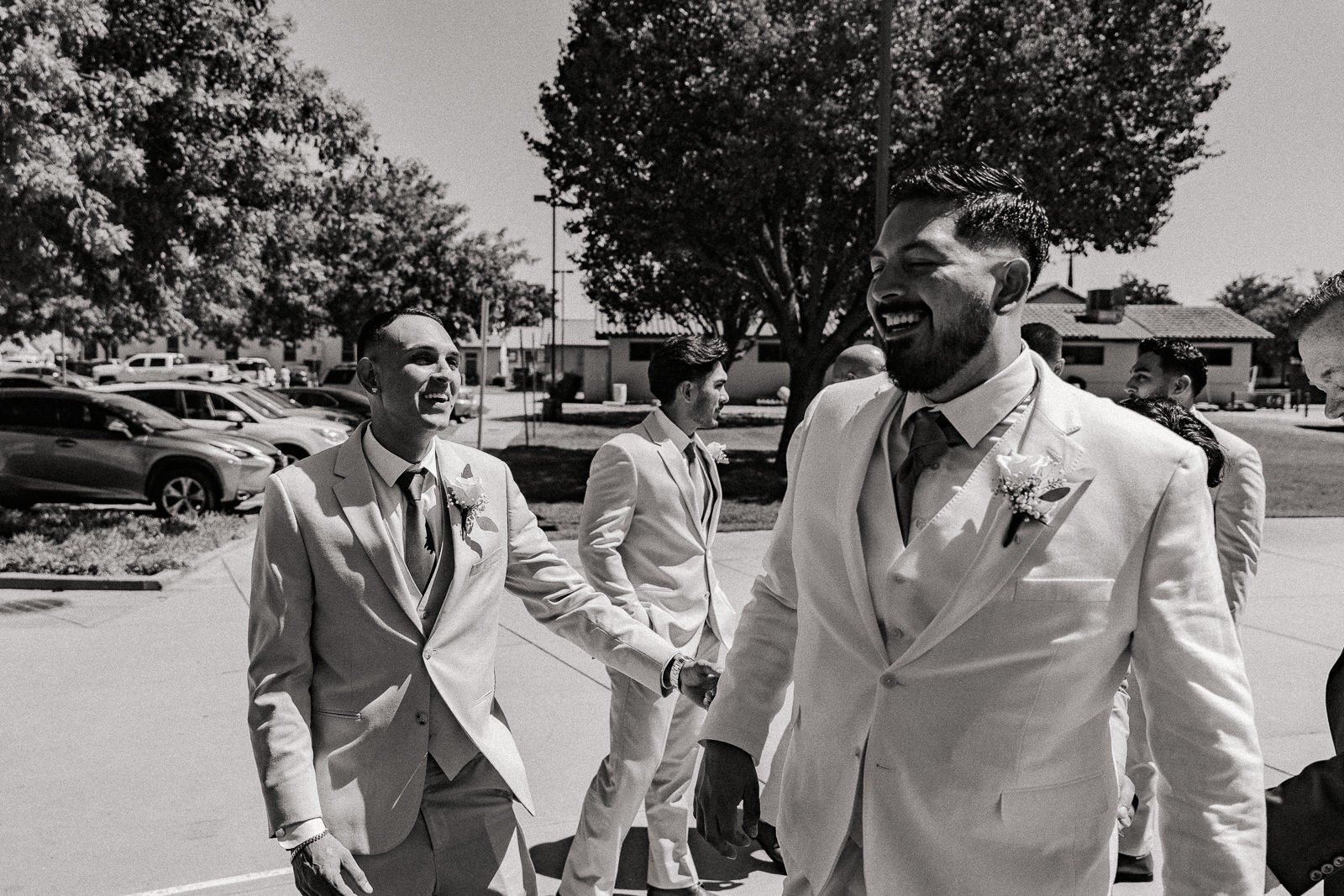 Groomsmen in light-colored suits laughing and interacting outdoors, with parked cars and trees in the background, in a black and white photograph.