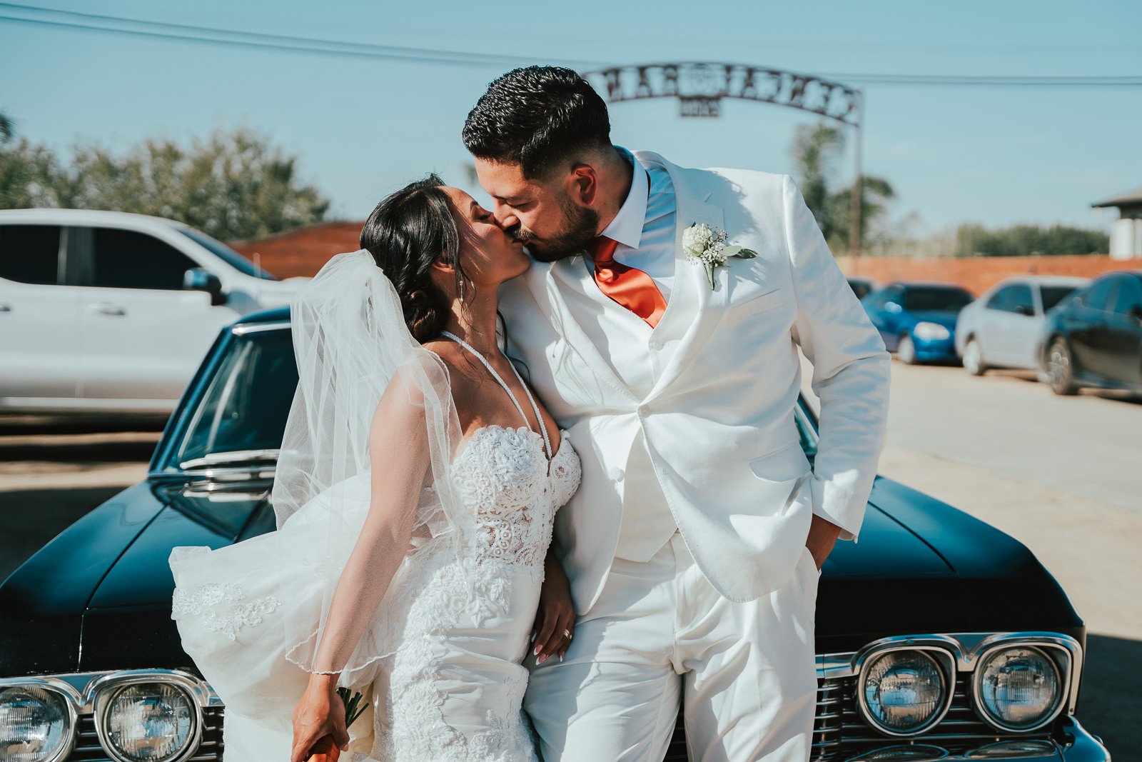 Bride and groom kissing in front of a black vintage car, bride in white lace dress and veil, groom in white suit with orange tie, outdoor setting with clear sky and parked cars.