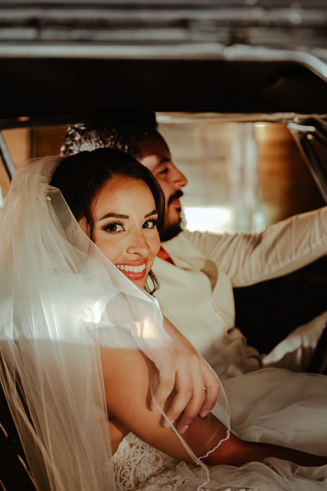 Bride and groom smiling inside a car, bride wearing a veil and white dress.