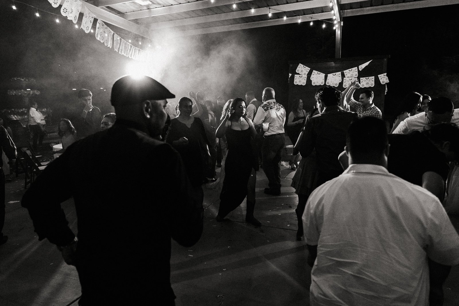 Black and white photo of people dancing at a lively party under string lights and papel picado decorations.