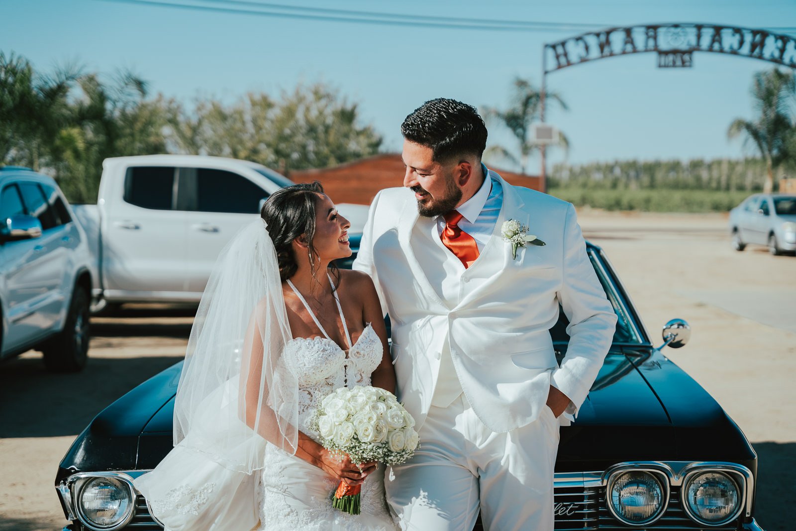 Bride and groom smiling, standing in front of a classic black car, with the bride holding a bouquet and both dressed in white wedding attire.