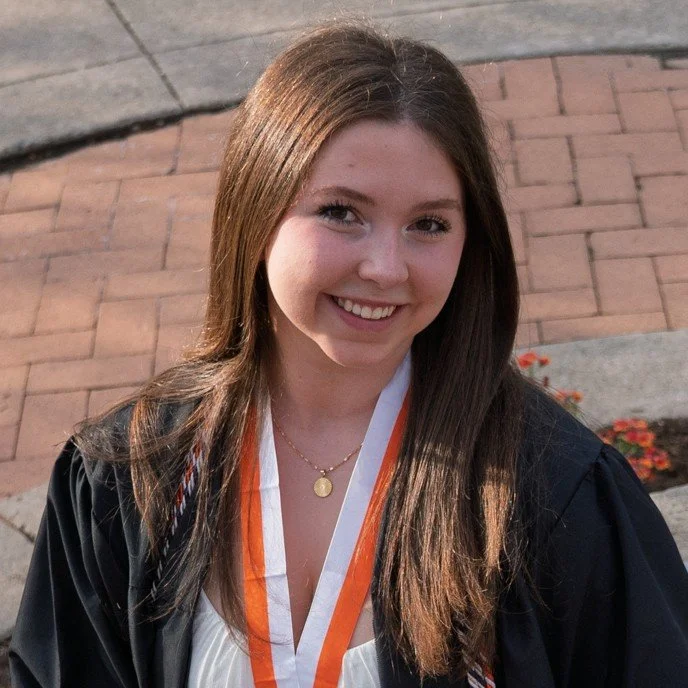 Young woman in graduation gown and medal, smiling outdoors on brick walkway.