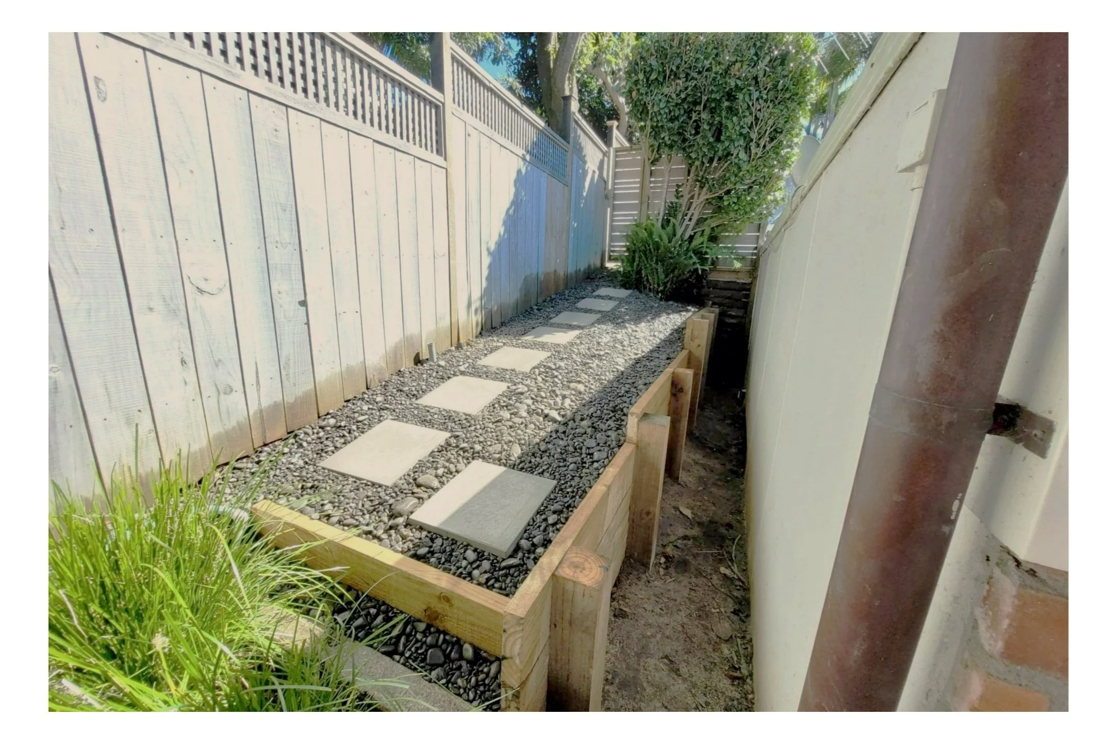 A narrow backyard pathway with square stepping stones on a gravel bed, on a built retaining wall.