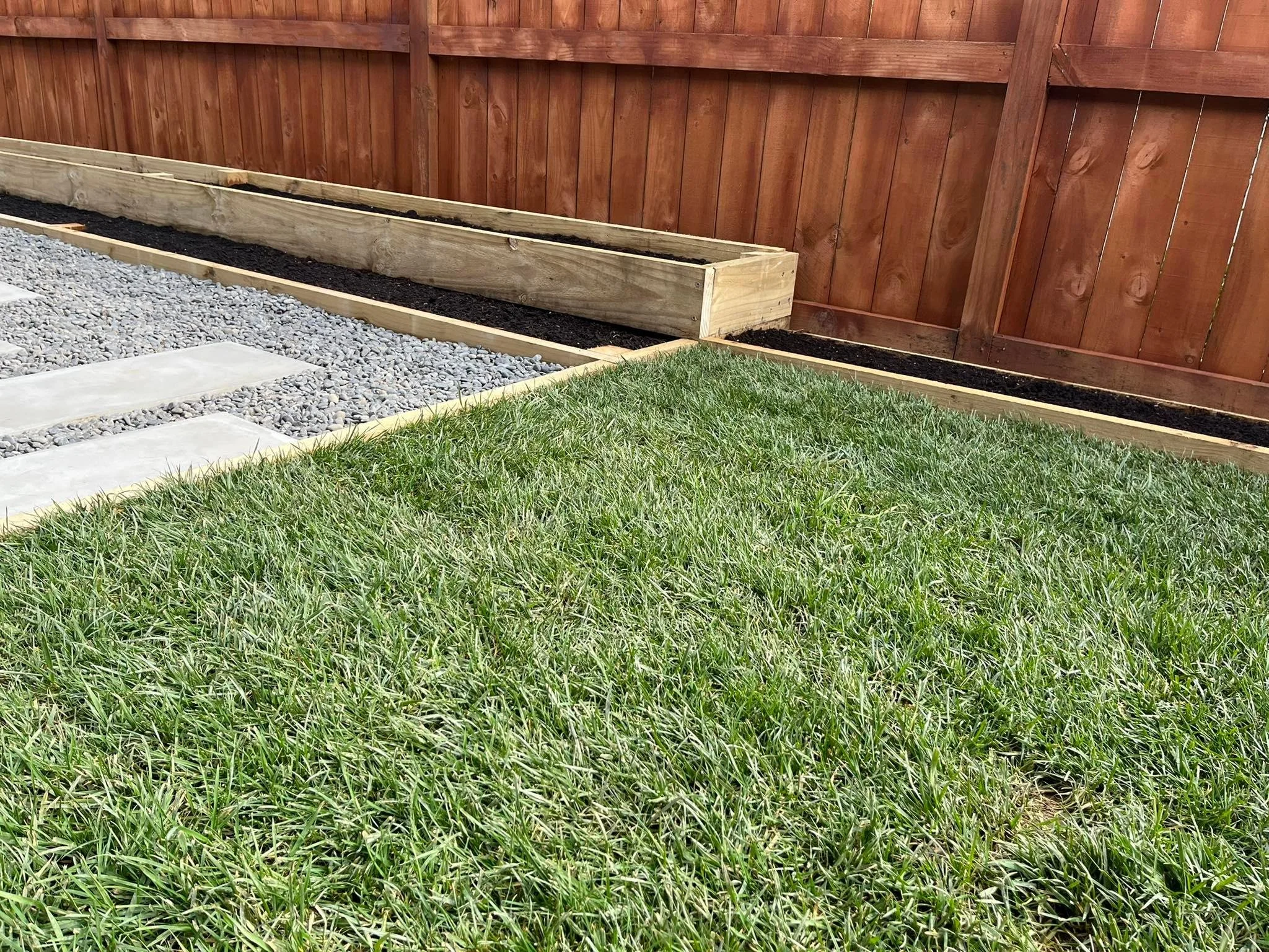 A backyard garden area with freshly laid wooden garden beds, gravel pathways, and a green lush ready lawn.