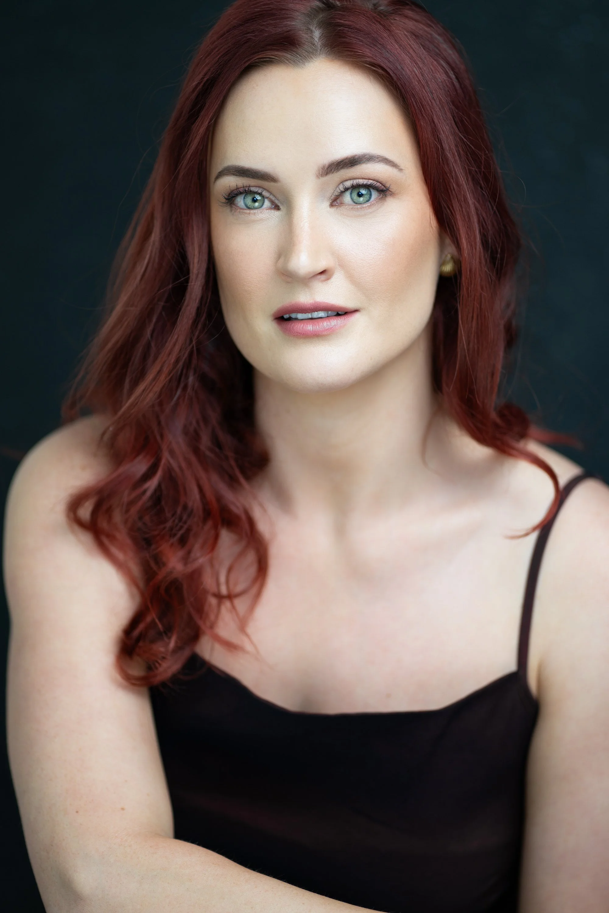 Professional headshot of Annie Aitken, a woman with long auburn hair and green eyes, wearing a black spaghetti-strap top. She gazes directly at the camera with a soft expression against a dark background.