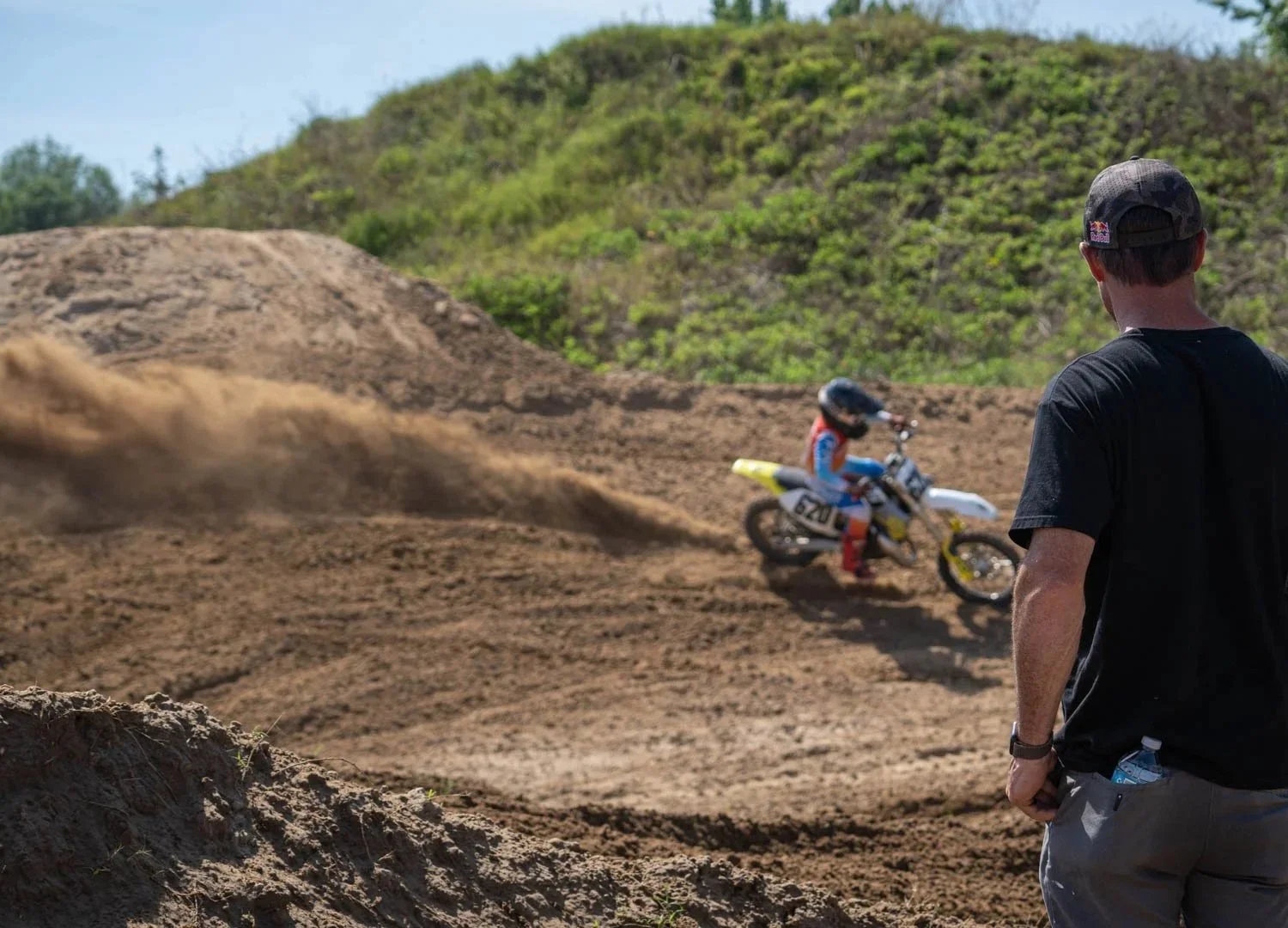 A man watching a young dirt bike rider go down a dirt track with hills and greenery in the background.