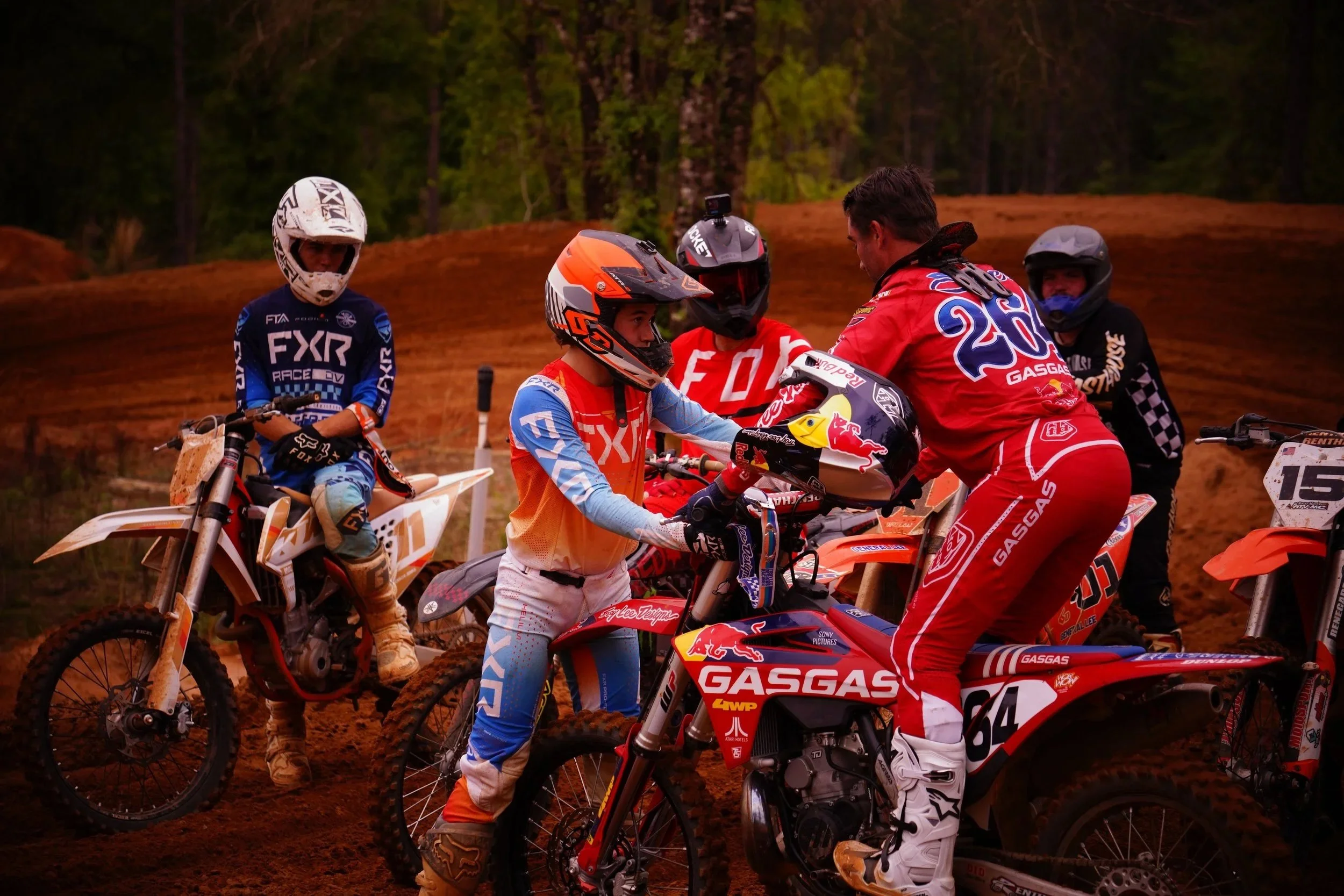 Motocross racers and a coach at a dirt track, with trees in the background.