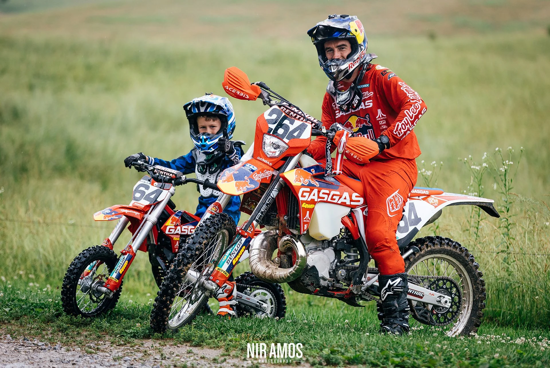 A man and a young boy with motorcycle helmets sitting on red and white KTM dirt bikes, on a grassy field.