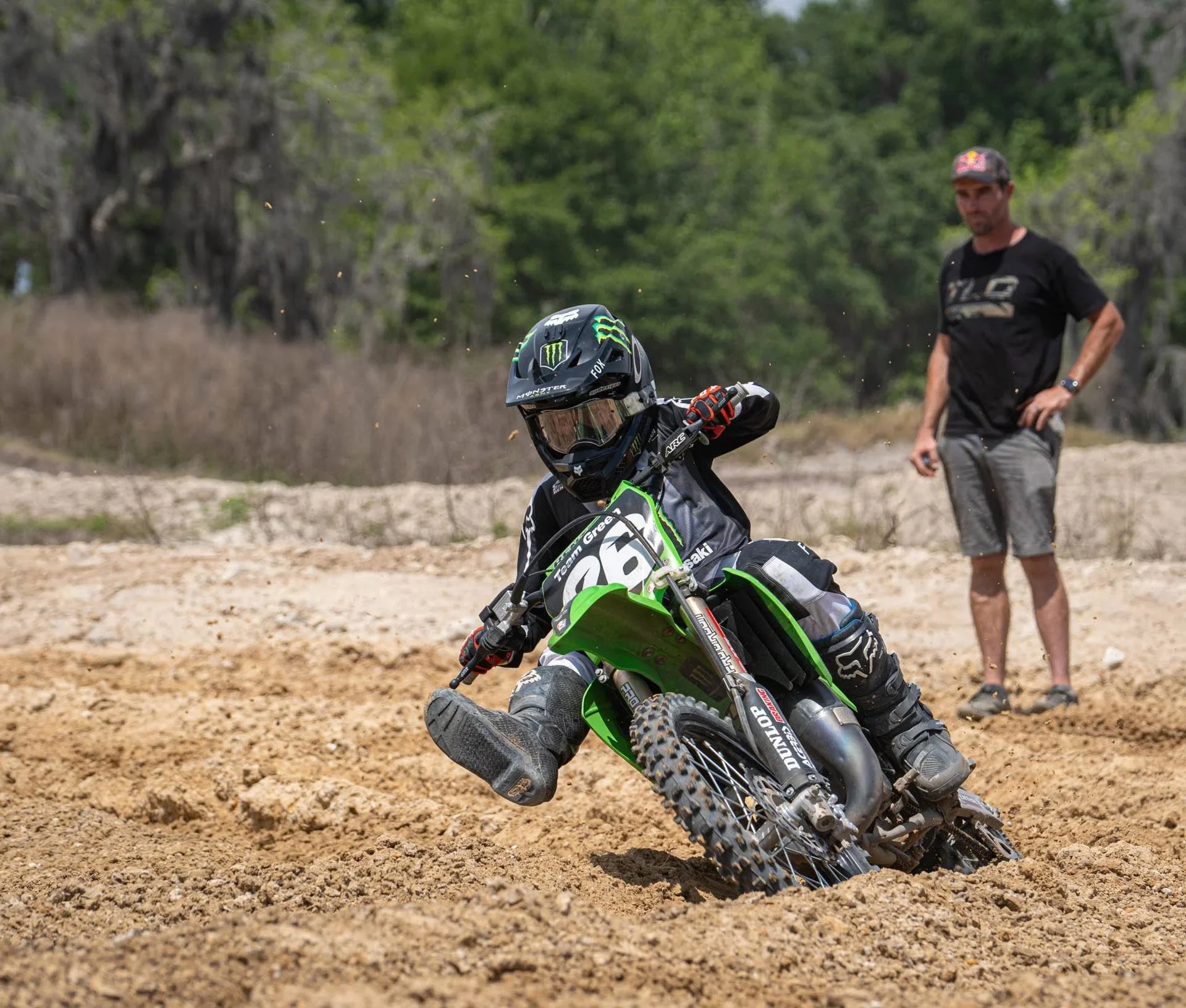 A person riding a green dirt bike on a sandy track, wearing a helmet and protective gear. An adult man is standing nearby watching.