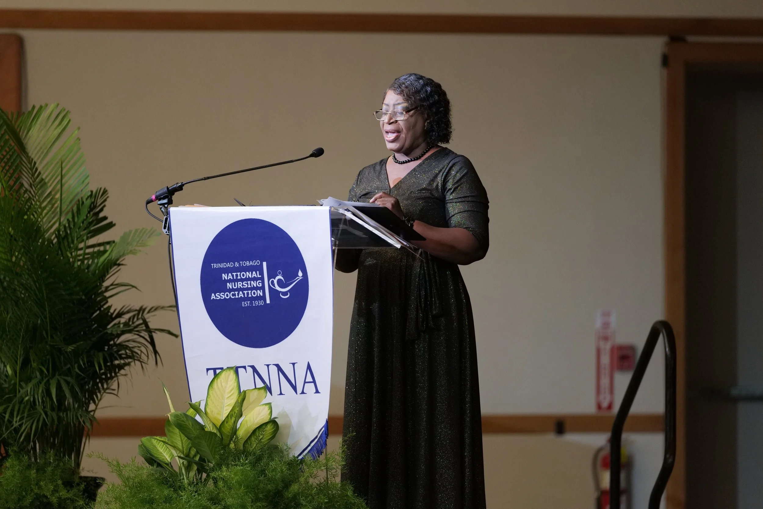 A woman in a black dress speaking at a podium with a Trinidad & Tobago National Nursing Association banner, surrounded by green plants, in a conference room.