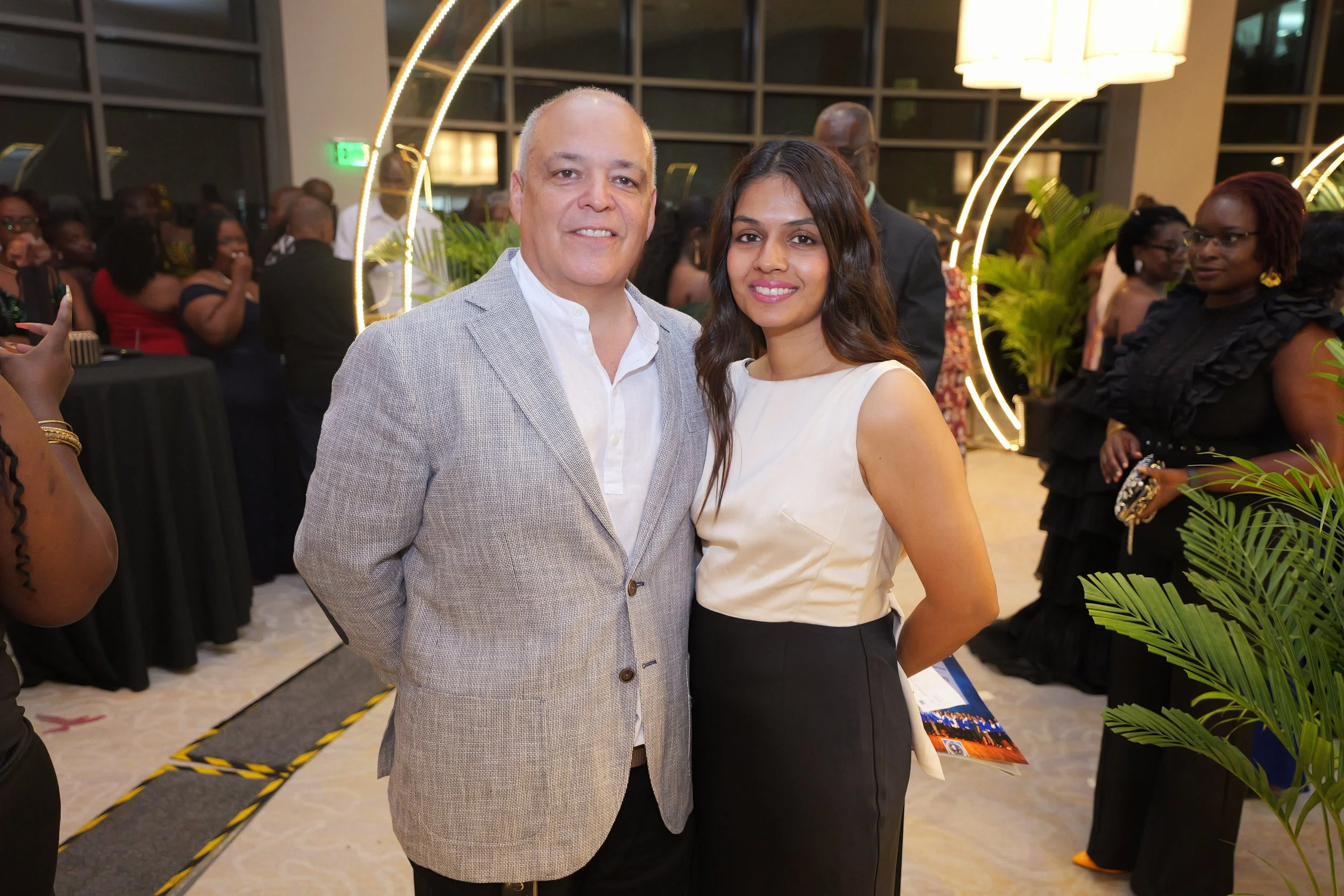 A man in a light gray blazer and a woman in a cream-colored top with a black skirt posing together at a formal event with other guests in the background.