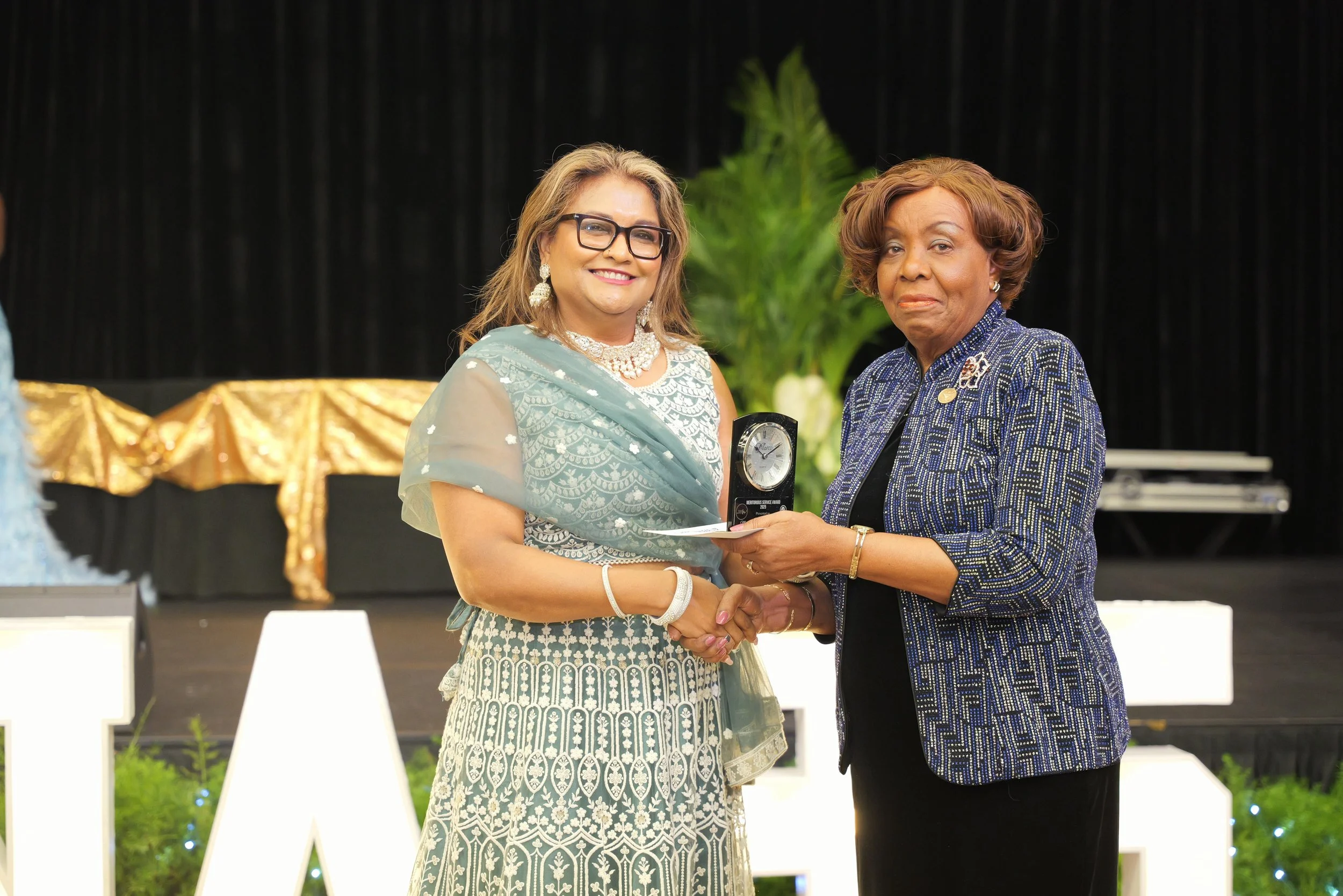 Two women shaking hands, with one woman presenting an award or trophy to the other, on a stage with black curtains and floral decorations.