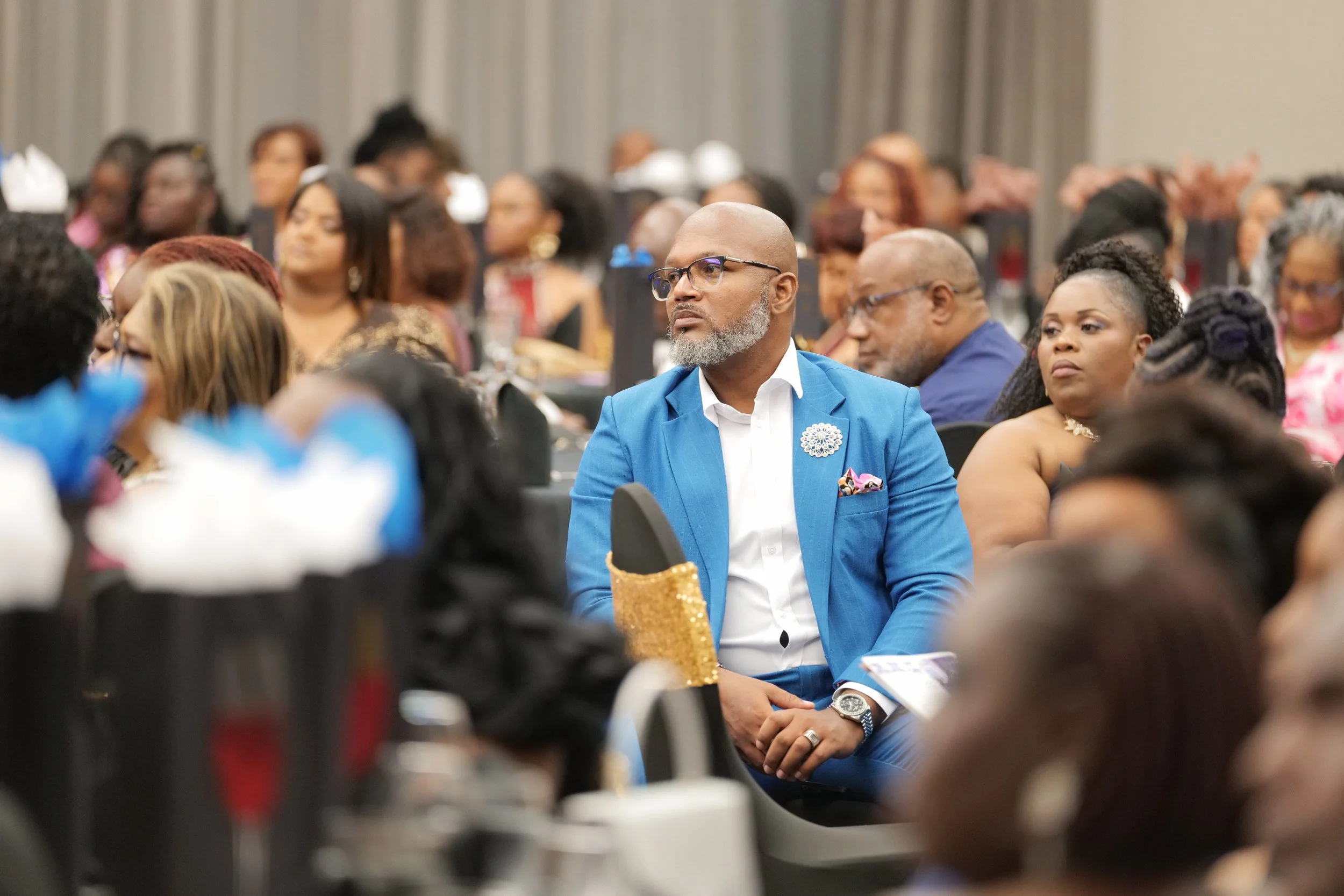 Crowd of diverse people attending an indoor event, with a man in a blue suit and glasses sitting attentively in the foreground.
