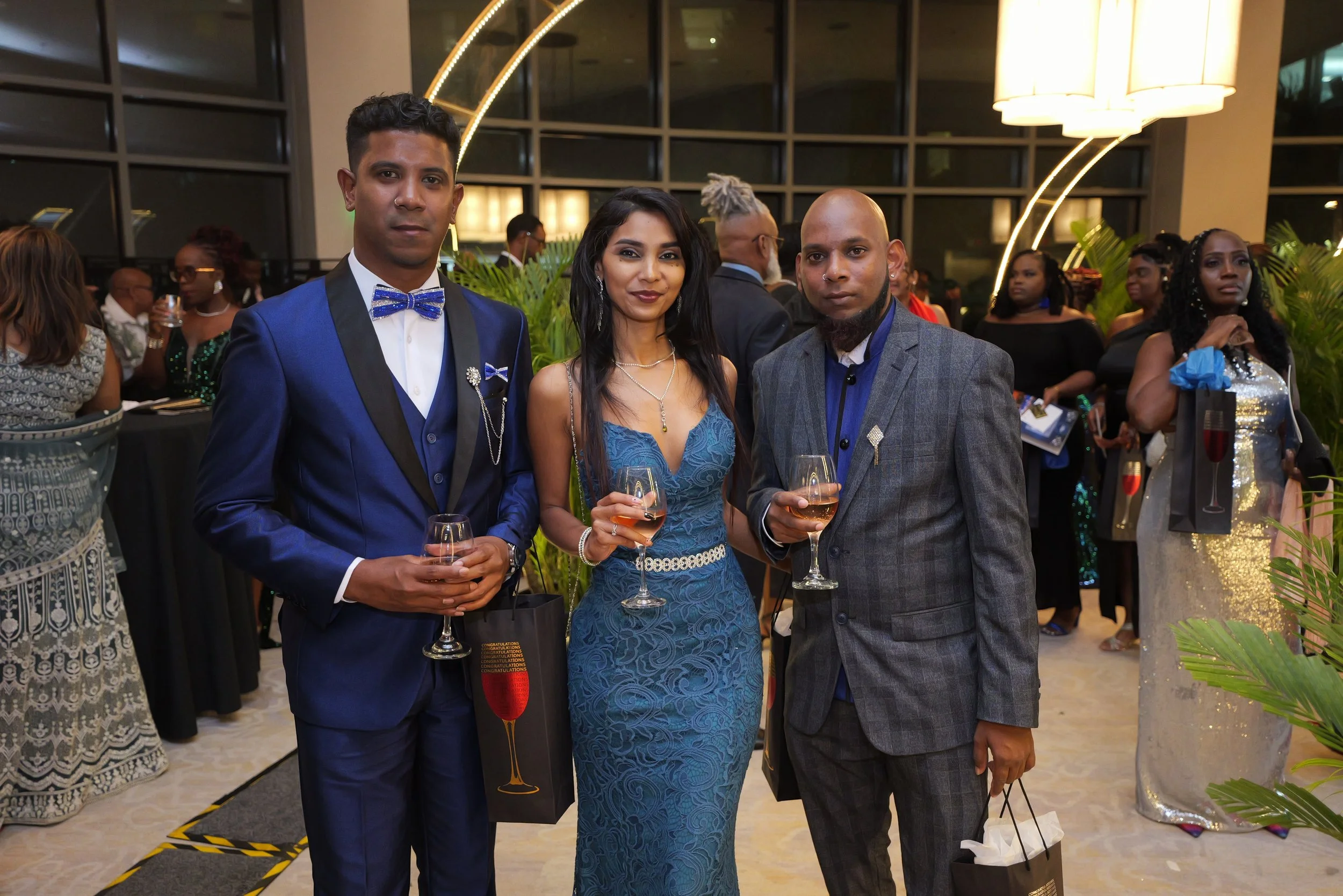 Three people dressed in formal attire standing together at a social event, holding glasses of wine. The man on the left is wearing a blue tuxedo with a black lapel and a blue bow tie. The woman in the center has long black hair, a blue lace dress, an