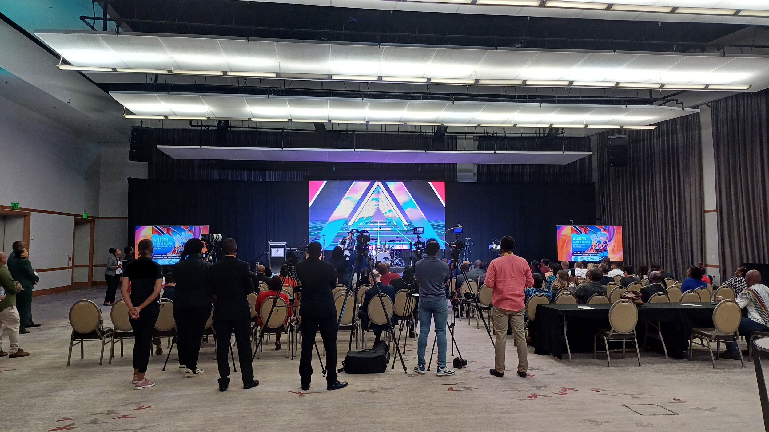 Conference room with a stage, large screens, and an audience, while media personnel stand in front taking photos and videos.