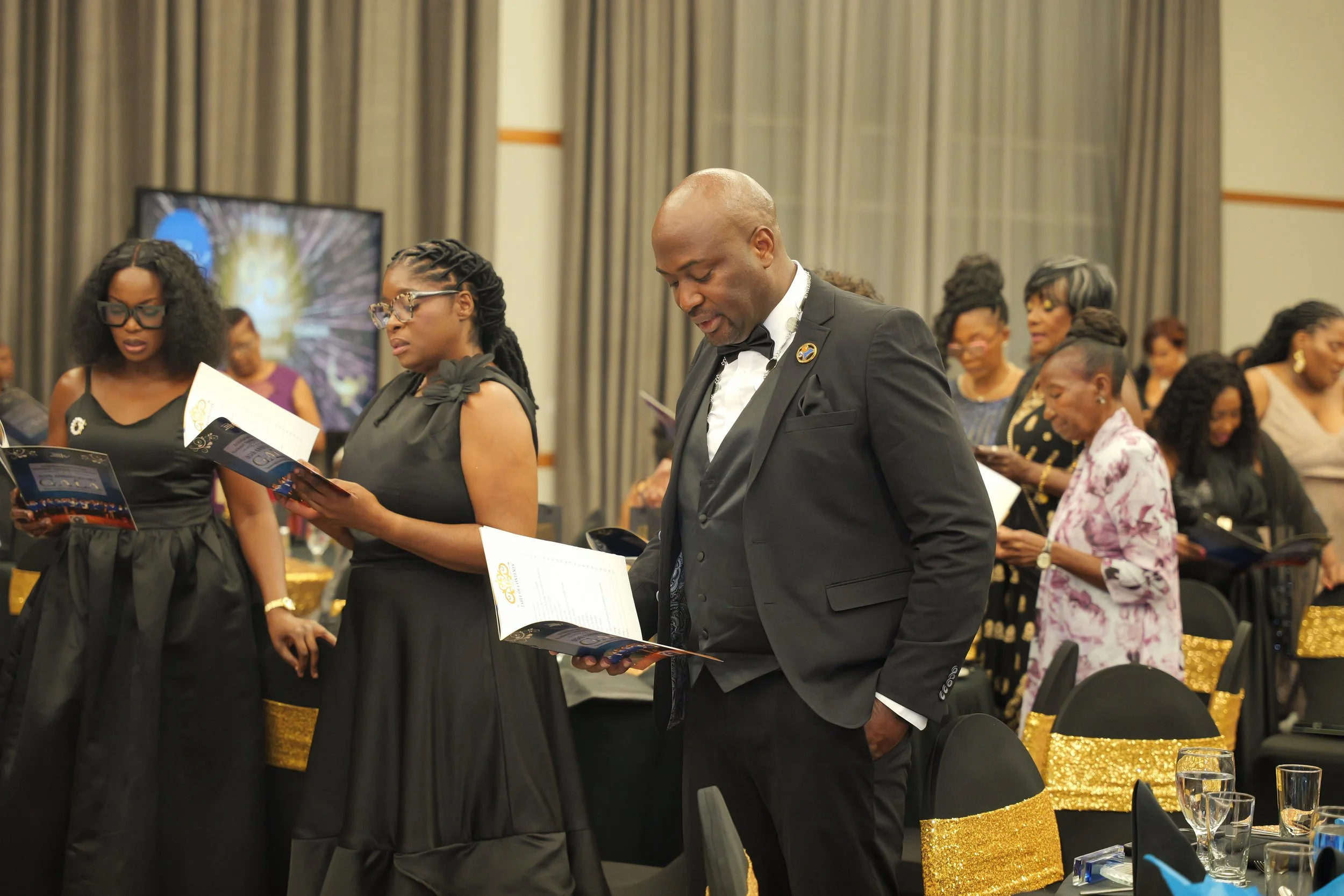 A group of people in formal attire, standing and reading programs at a formal indoor event, with gold and black decor.