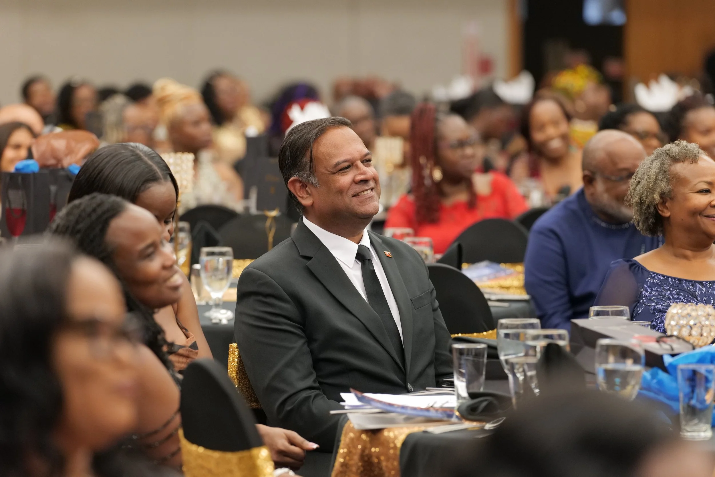 A man in a black suit and tie is sitting at a formal event, smiling and looking to his right. He is surrounded by other people who are also seated at tables, dressed in formal attire.