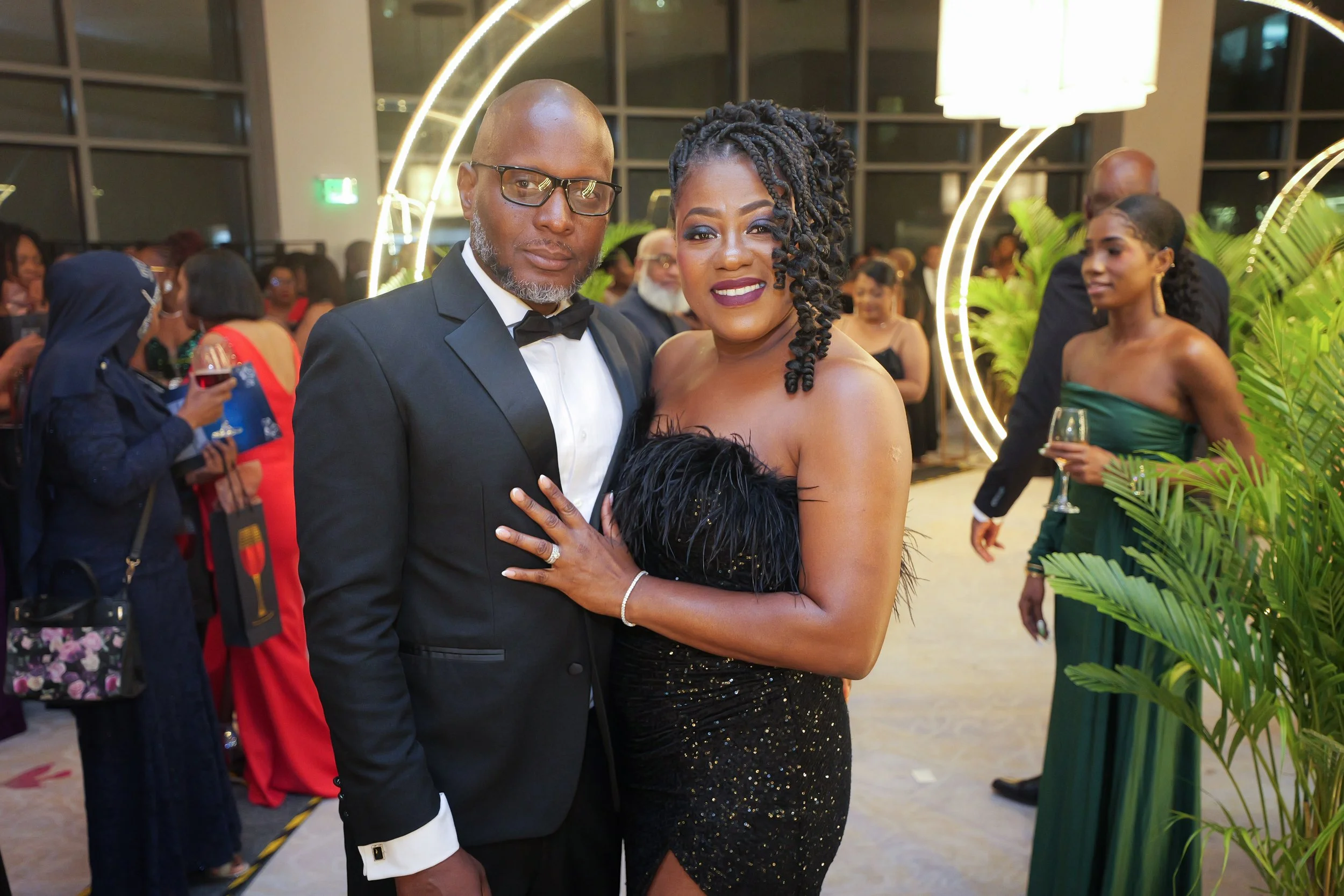 A couple dressed in formal attire at a celebration or wedding. The man is wearing a black tuxedo with a bow tie and glasses. The woman is wearing a black, sparkly strapless dress with feather details, and has styled her hair in twists. They are stand