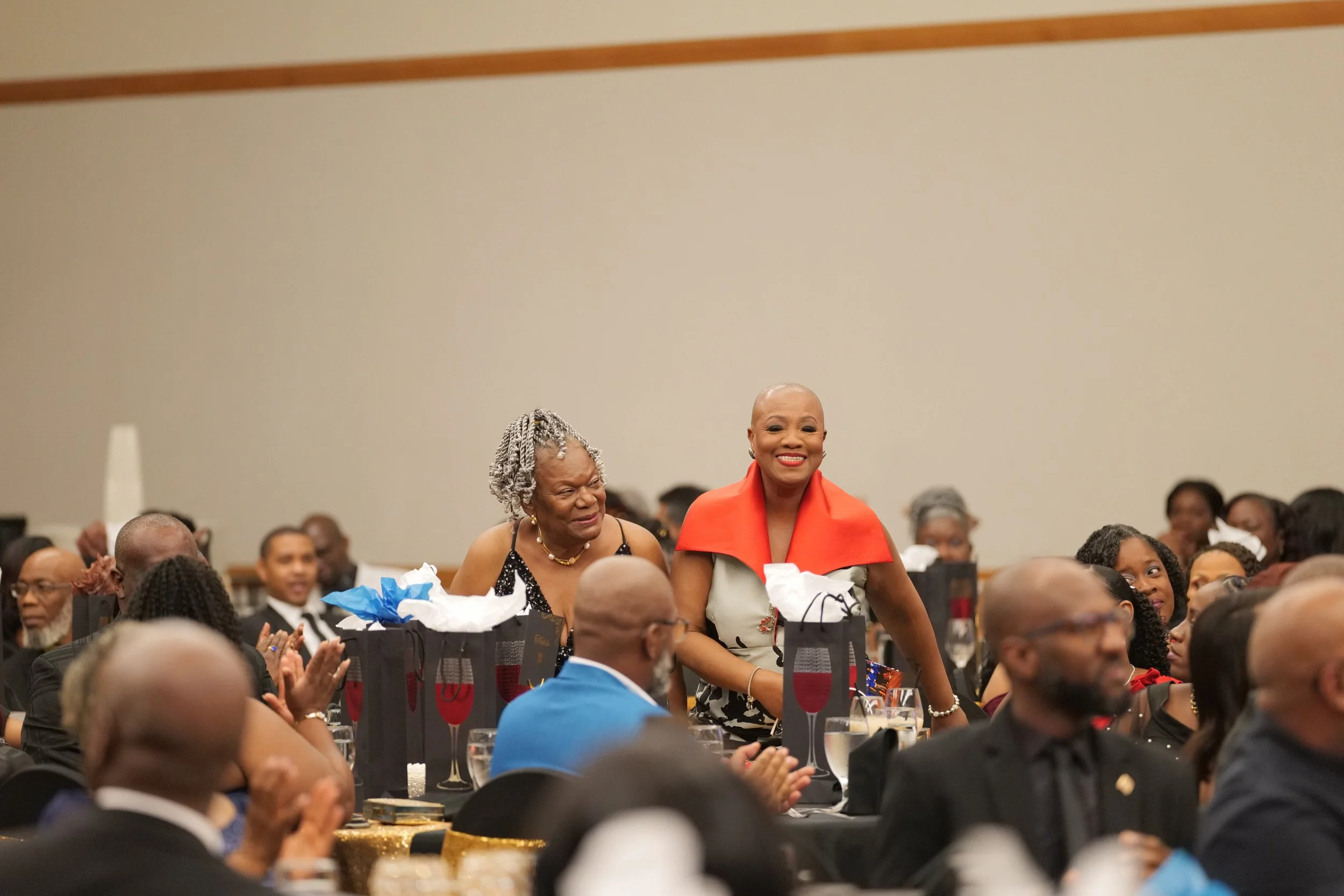 A group of people attending a formal event, with two women standing and smiling amidst seated guests. The woman on the right has a bald head and is wearing a beige and red outfit, while the woman on the left has gray curly hair and is dressed in blac