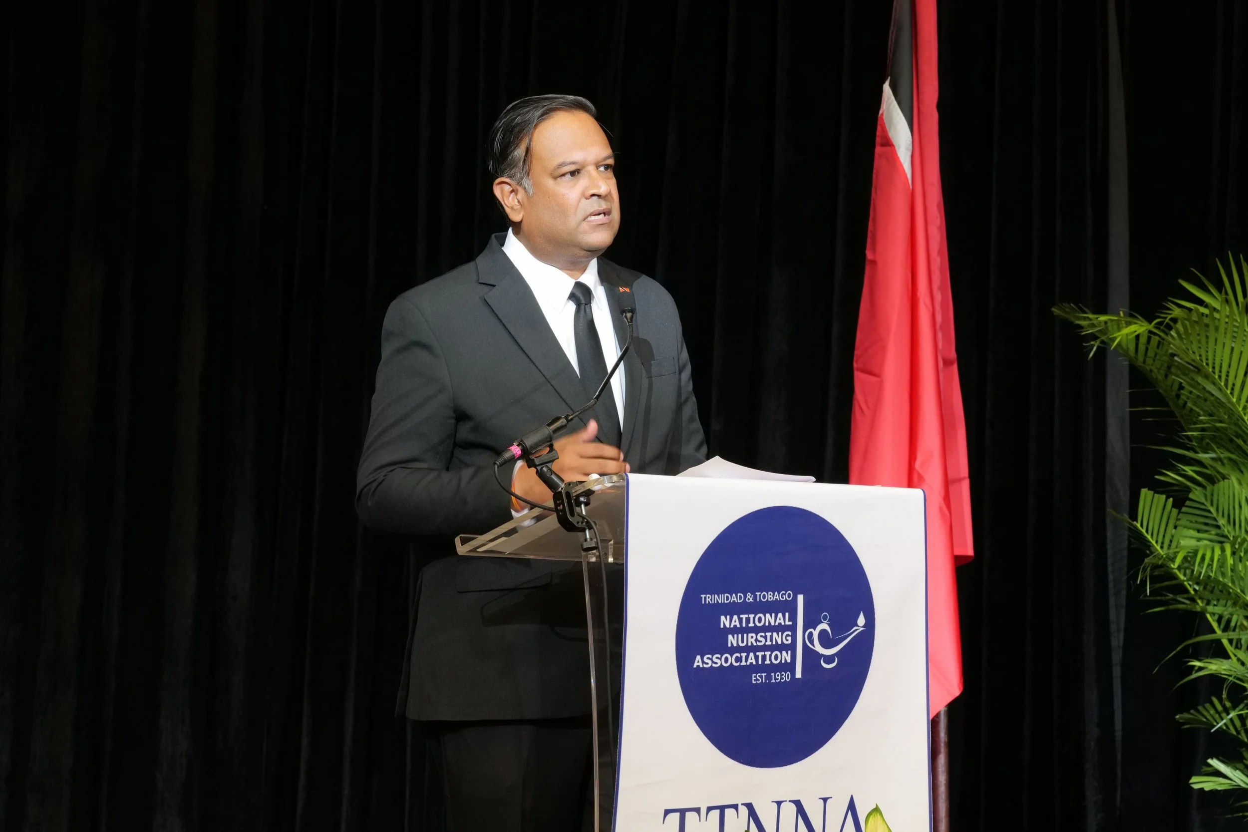 Man in black suit and tie speaking at a podium decorated with the Trinidad and Tobago National Nursing Association logo, with a red and white flag and green plants in the background.