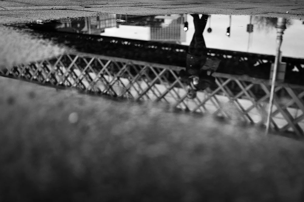 Reflection of a person walking near a bridge in a puddle on the ground, with a blurry foreground, Dublin City Street Photography.