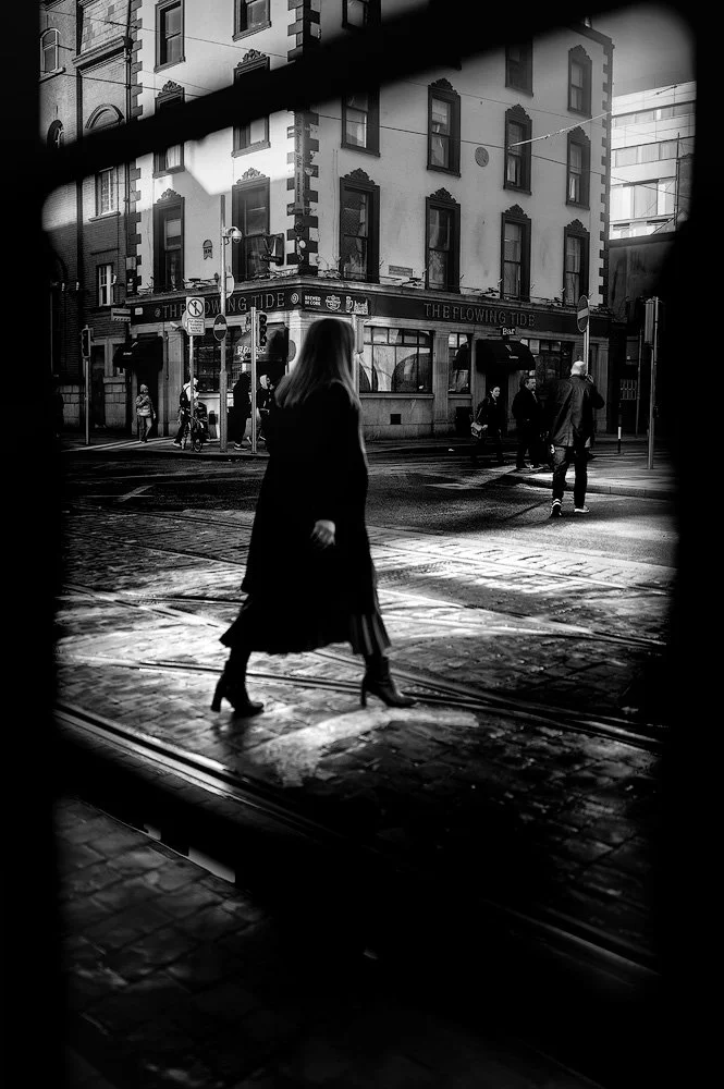 Black and white photo of a woman in a long coat and heeled boots walking across a street in an urban area, with buildings, pedestrians, and tram tracks visible.