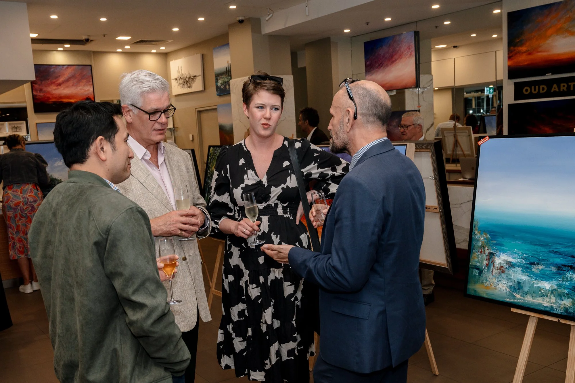 Group of four people conversing at an art gallery, holding glasses of champagne, with paintings displayed in the background.