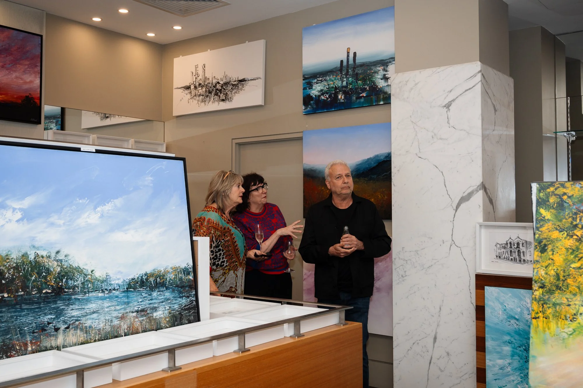 Three people viewing and discussing artwork in an art gallery, with paintings and photographs displayed on the walls and on stands around them.