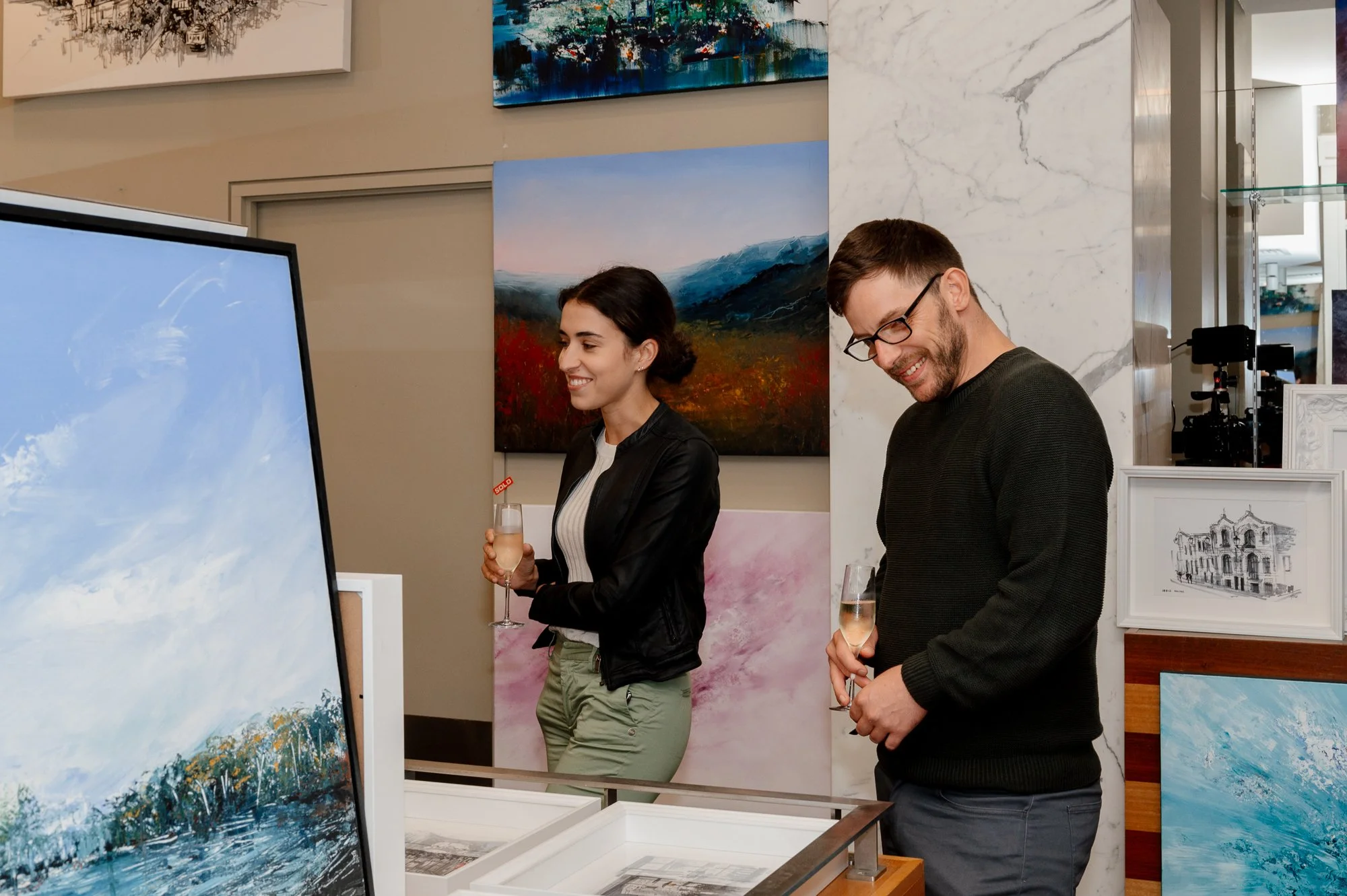 Two people viewing paintings at an art gallery, each holding a glass of champagne, surrounded by various colorful landscape artworks.