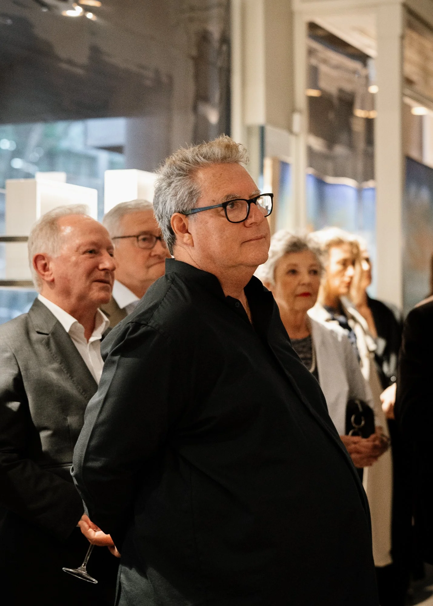 A group of people at a social event, with one man in the foreground wearing glasses and a dark jacket, attentively listening. Other guests, including an older woman and men, stand behind him in an indoor setting with large windows and modern decor.