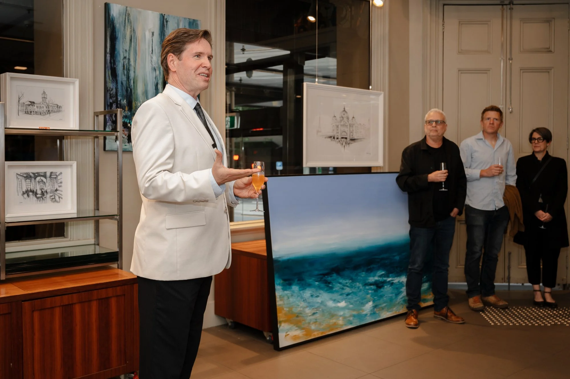 Man in a white suit giving a speech at an art gallery reception, holding a glass of champagne. Three people stand behind him, listening, in front of framed artwork and paintings.