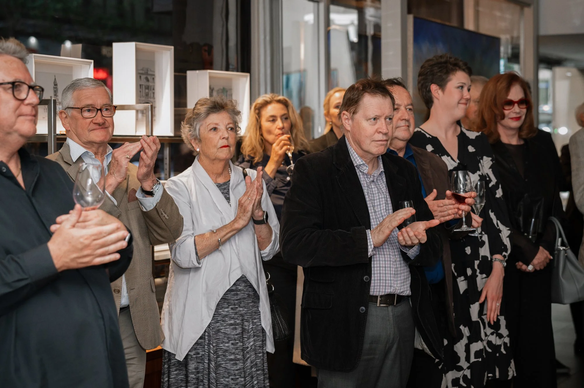 Group of people at a social event, standing indoors with some holding glasses of wine, attentively watching or listening.