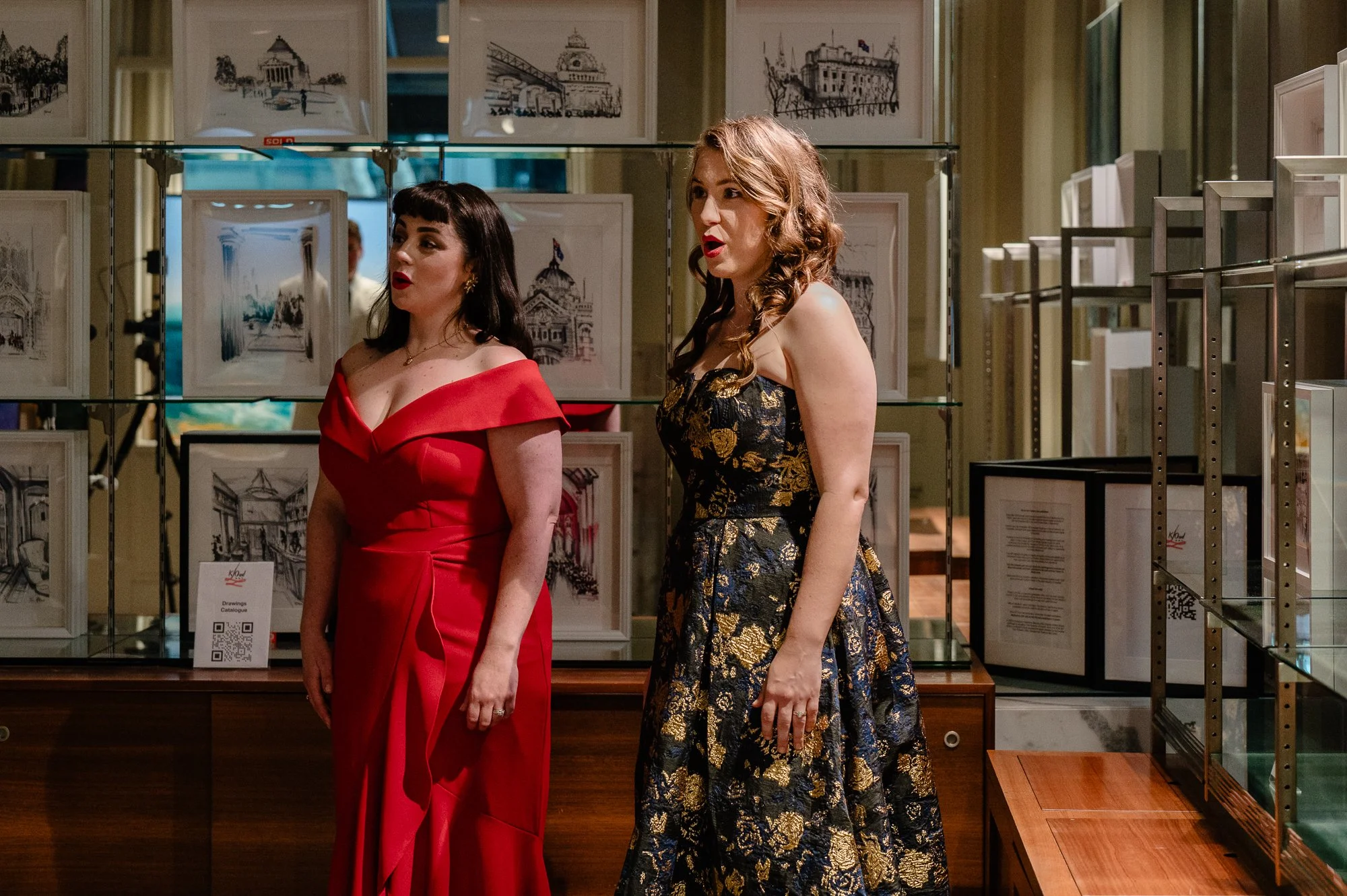 Two women in elegant dresses standing in an art gallery, looking at artwork displayed on glass shelves behind them.