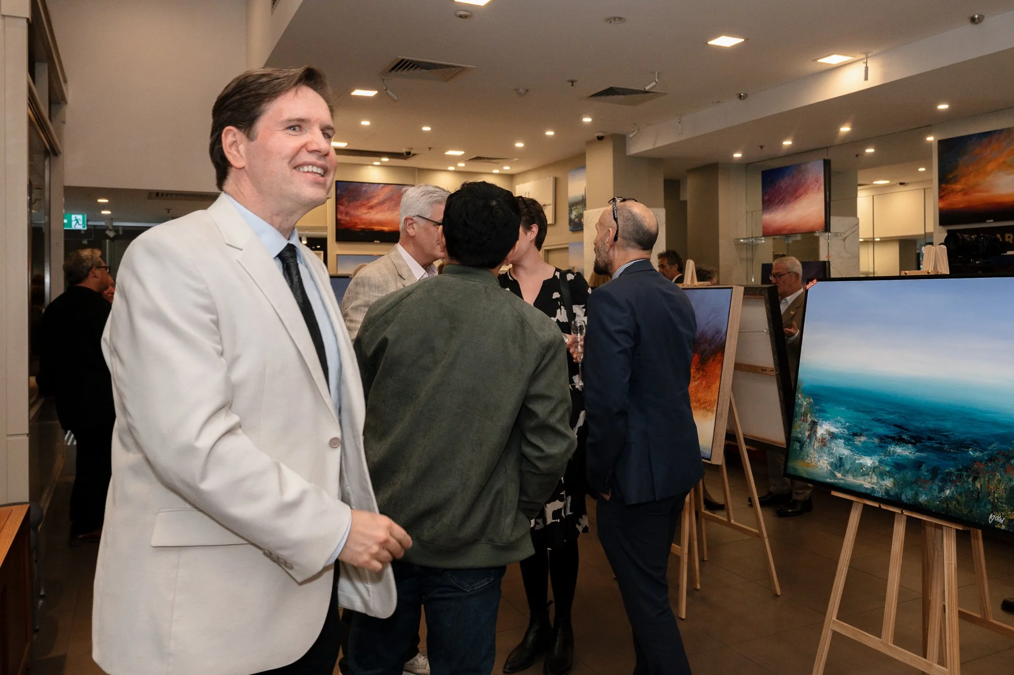 Group of people at an art gallery, viewing landscape paintings displayed on easels.