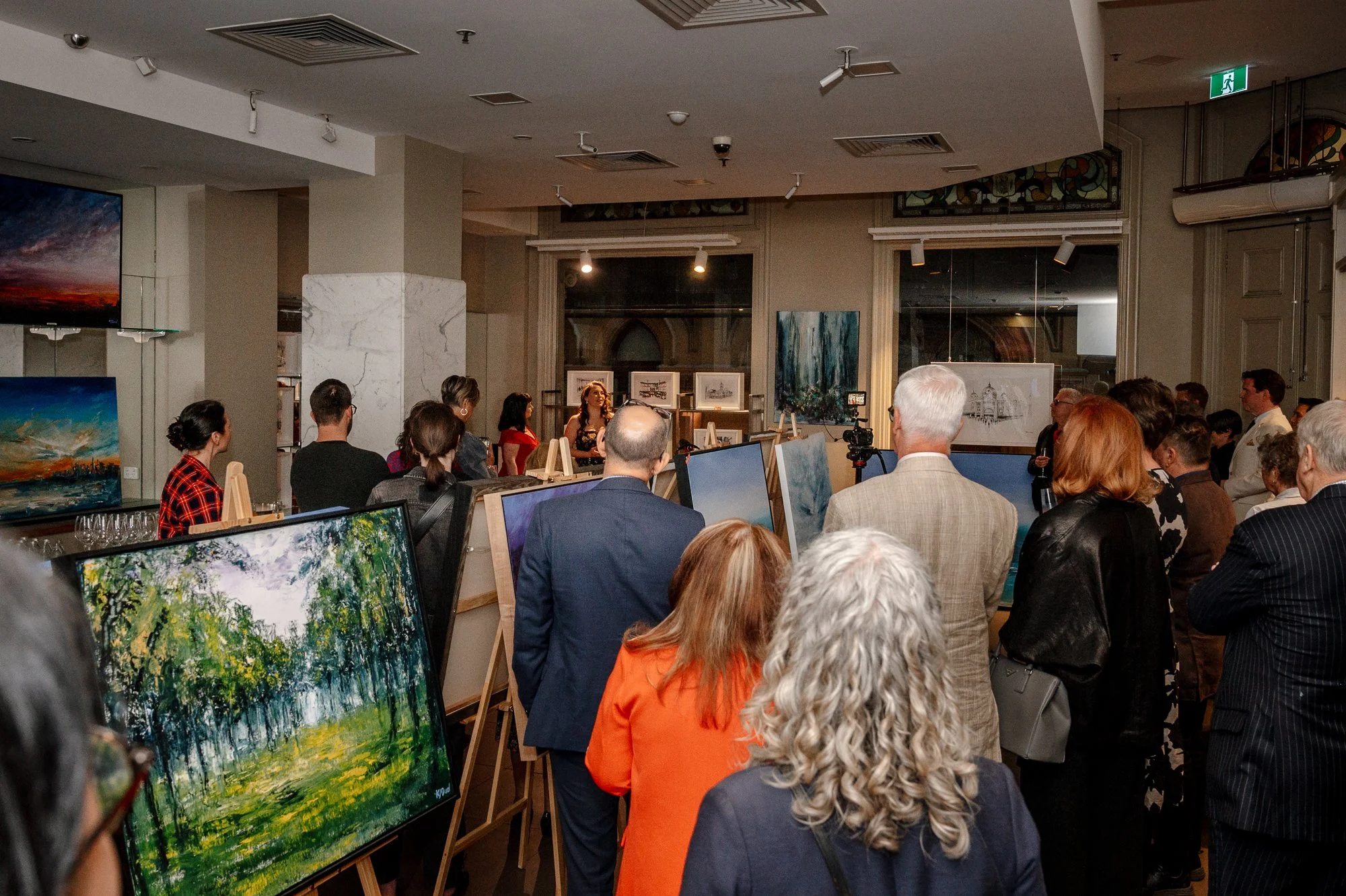 An art gallery event with people viewing paintings displayed on easels and walls, and a speaker or artist presenting in front of an audience.