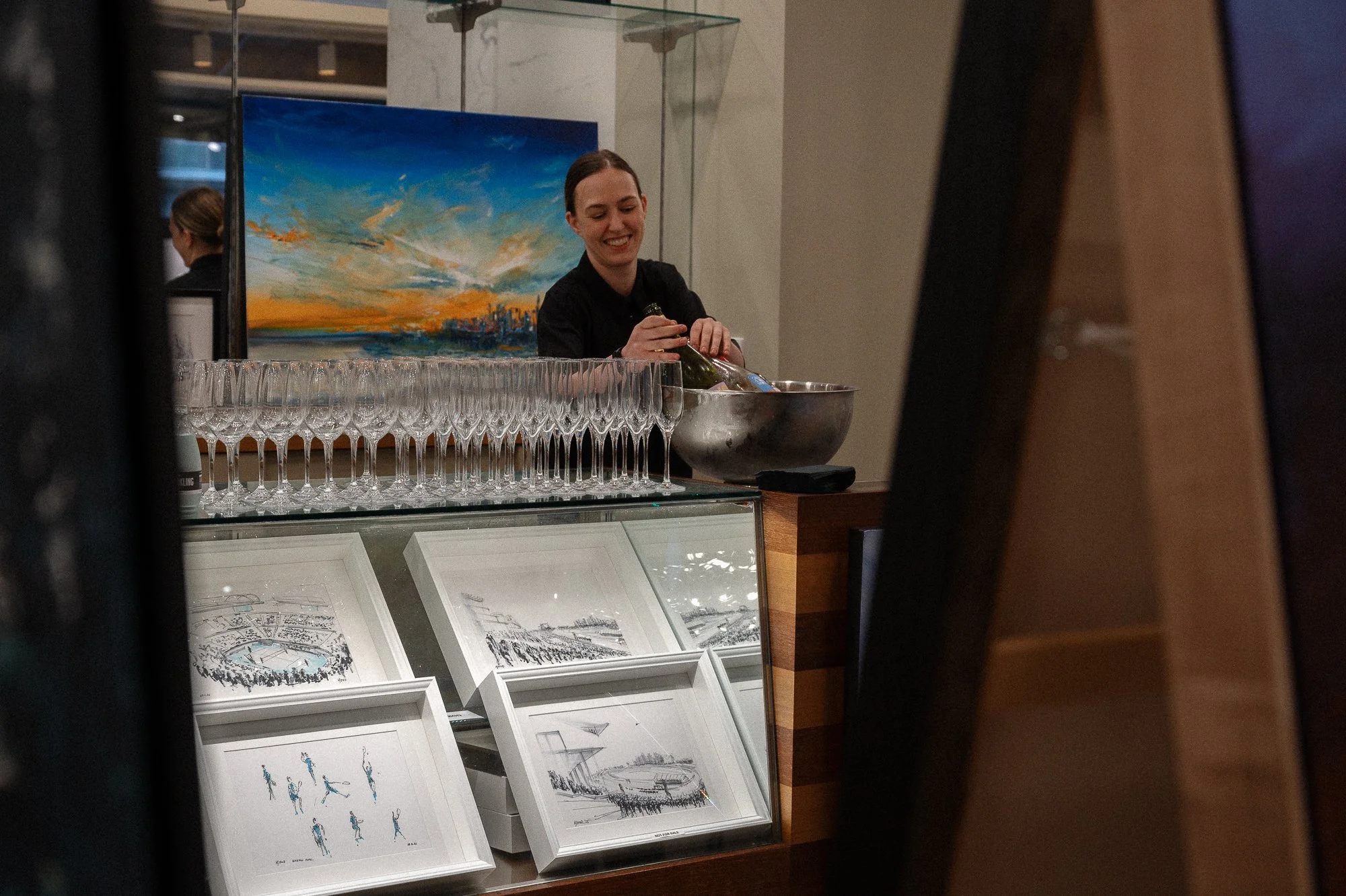 A woman preparing drinks behind a bar counter with wine glasses, framed artwork, and sketches of stadiums.