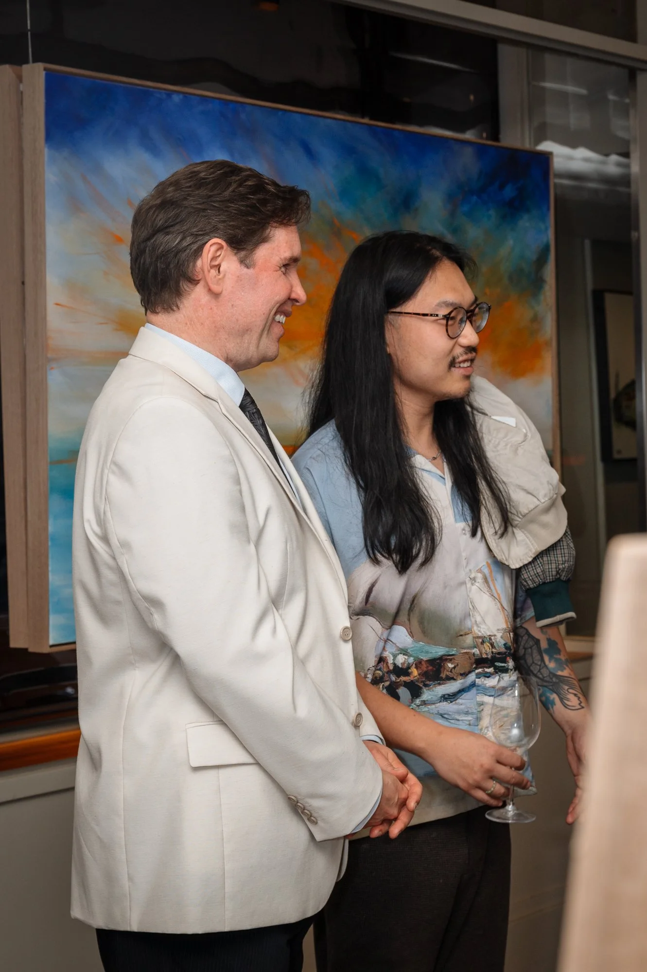 Two men standing and talking at an art gallery with a colorful painting in the background, one wearing a white suit and the other wearing glasses and a colorful shirt, holding a wine glass.