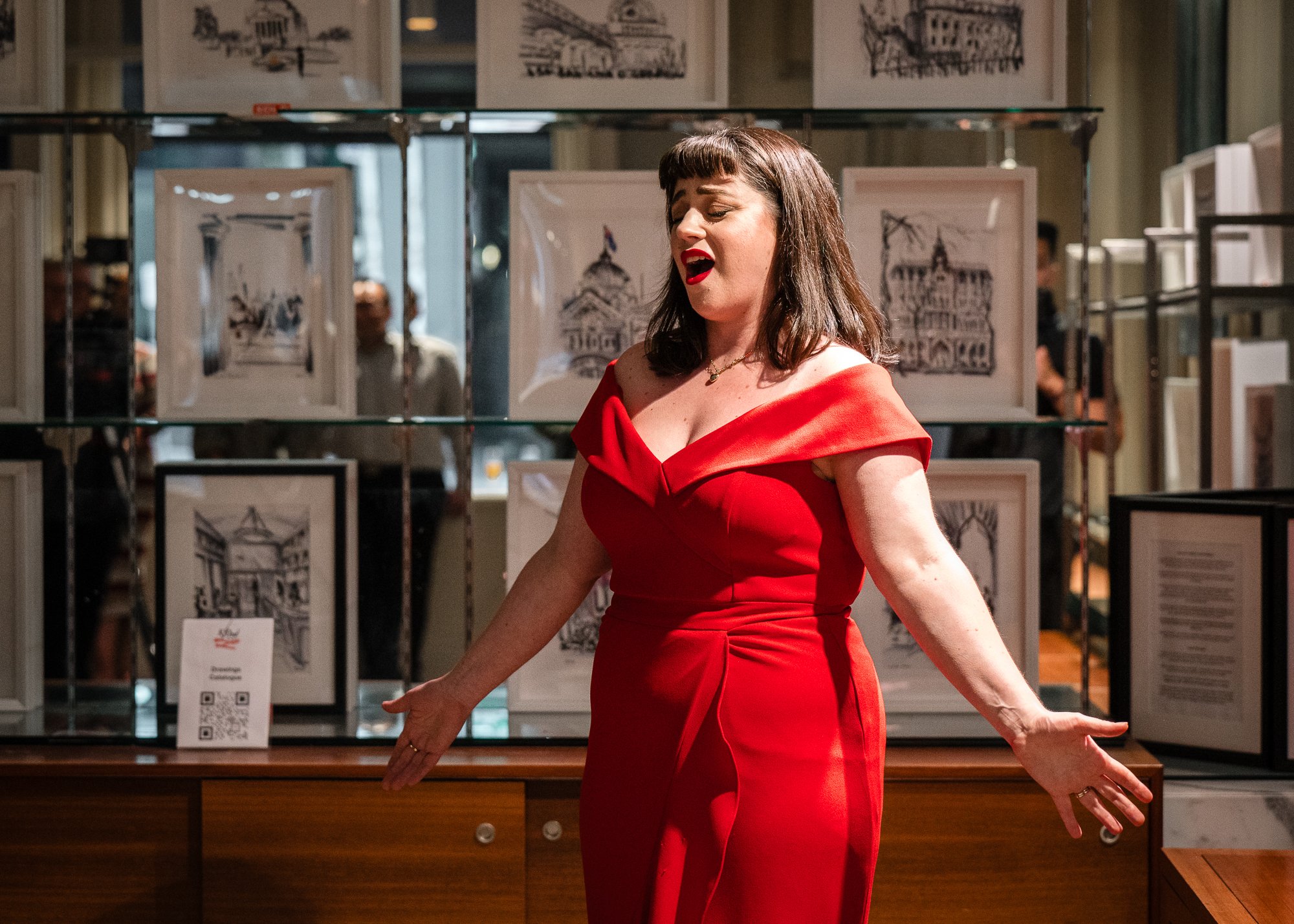 A woman wearing a red dress singing or performing in an indoor setting with framed artwork on display in the background.