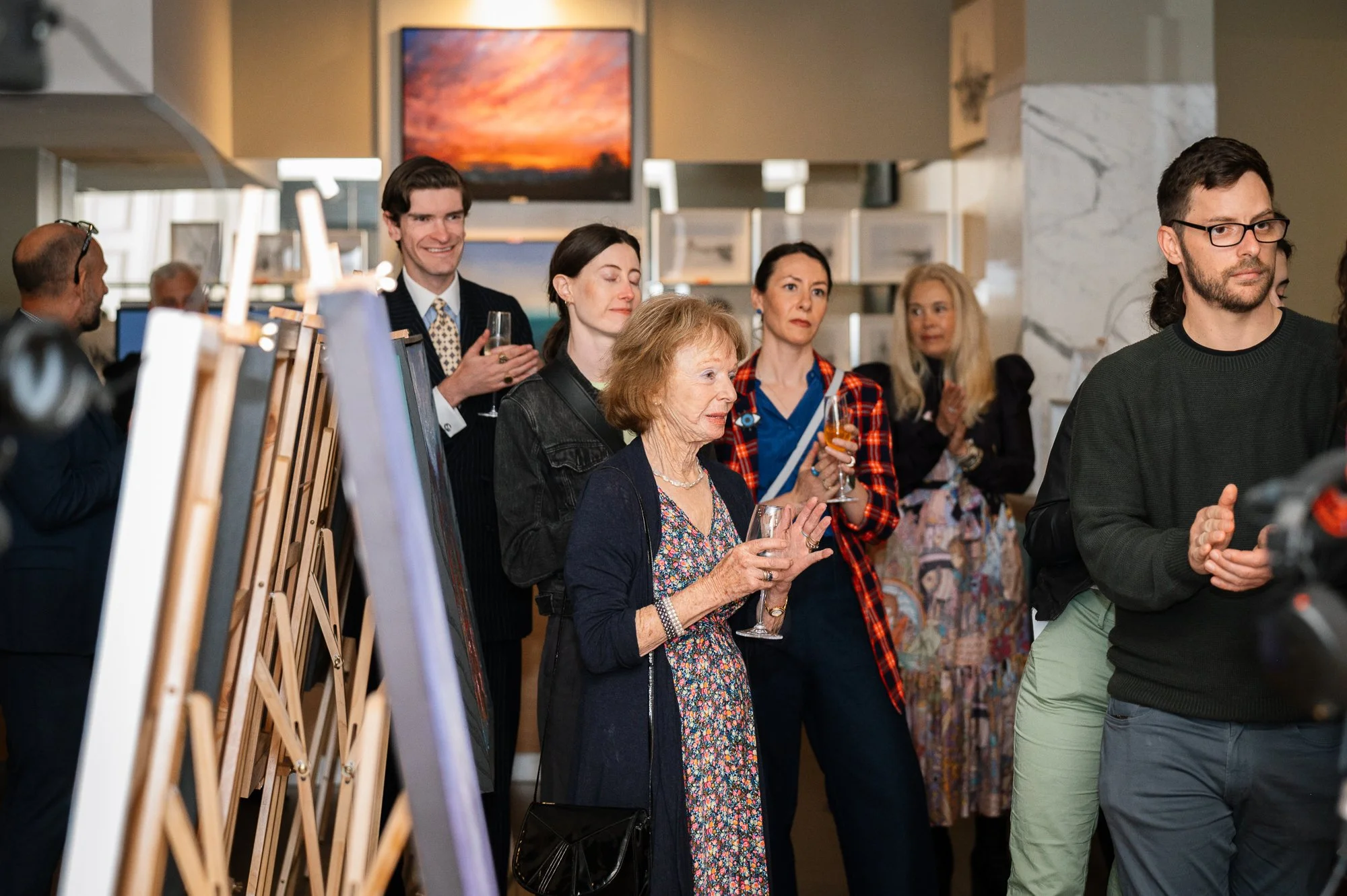 People attending an art gallery or exhibition, observing artwork displayed on easels, with some holding drinks and paying attention to the event.