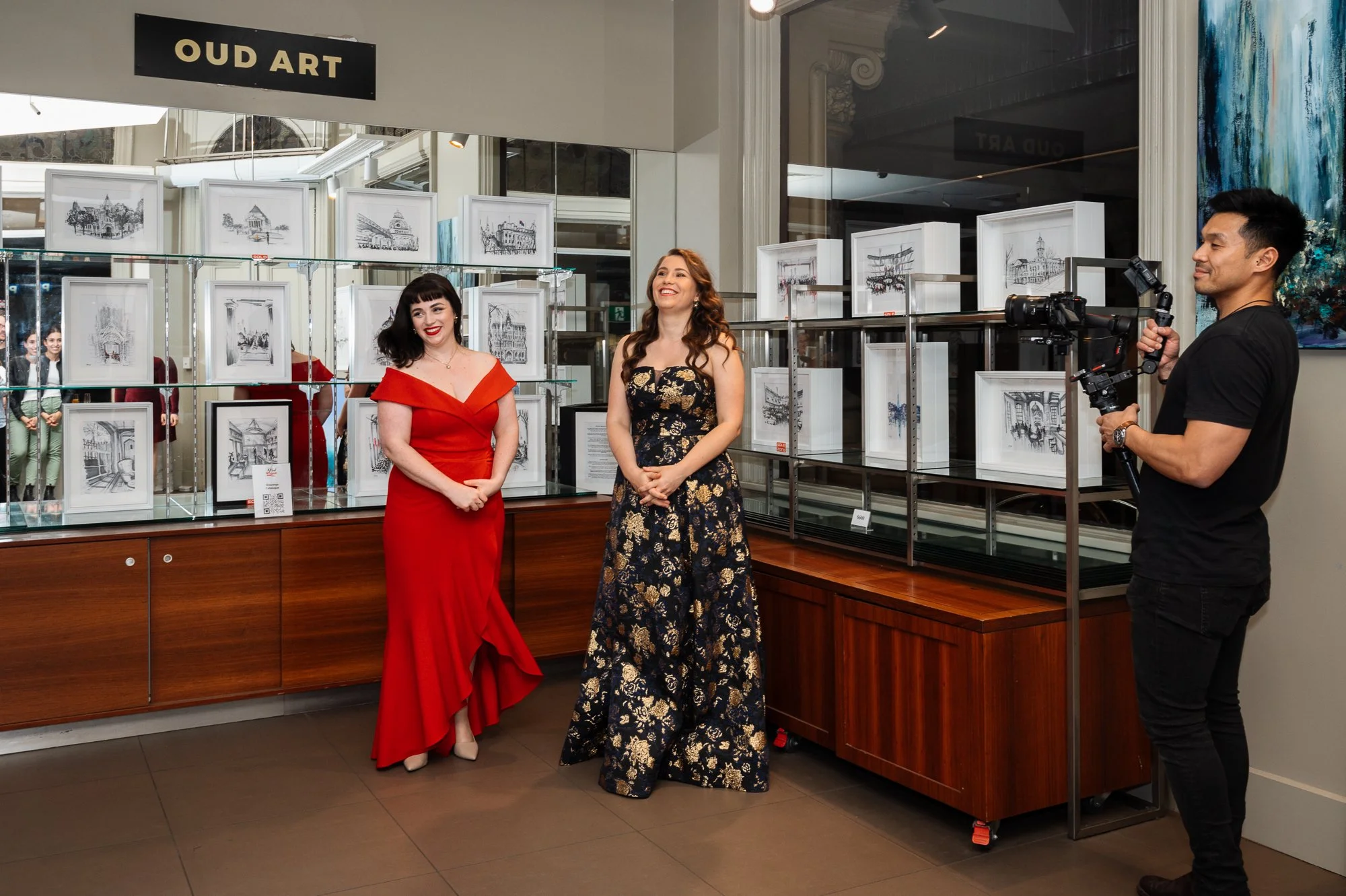 Two women in evening dresses are being filmed by a man with a camera at an art exhibit titled 'OUD ART' featuring framed artwork displayed behind glass.