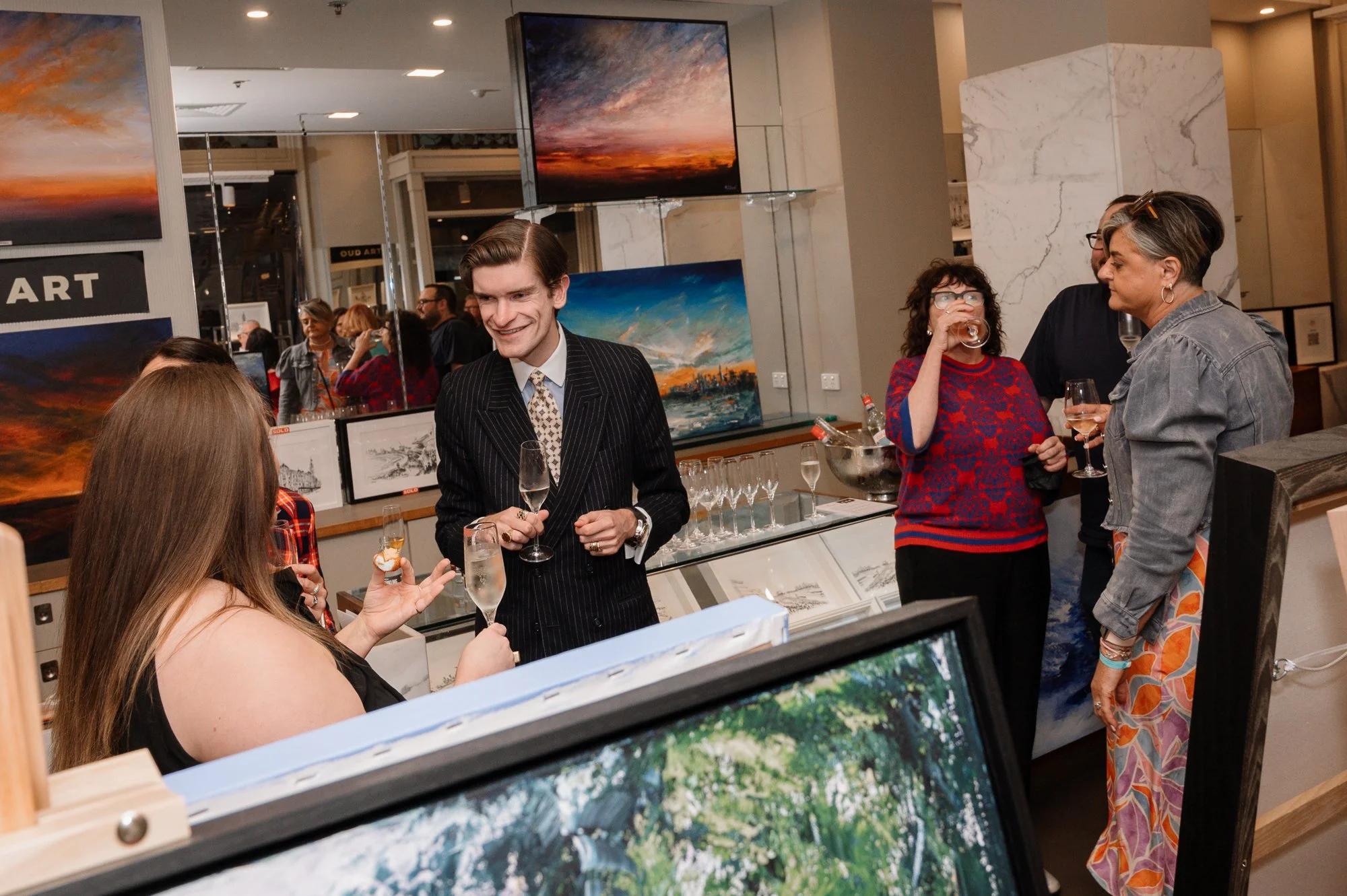 People at an art gallery engaging in conversation and holding drinks, with paintings displayed on the walls behind them.