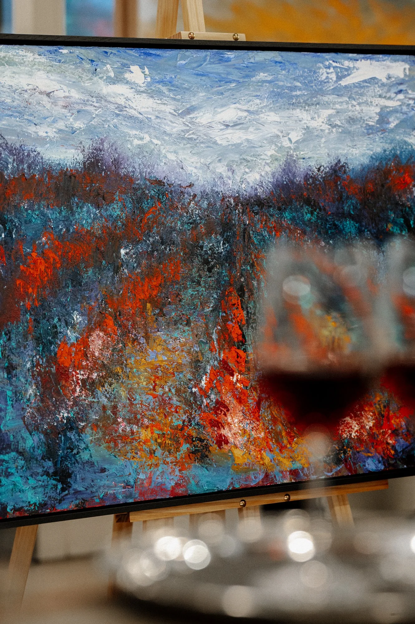 Close-up of an abstract landscape painting with winter mountains and colorful trees, displayed on an easel in an art gallery.