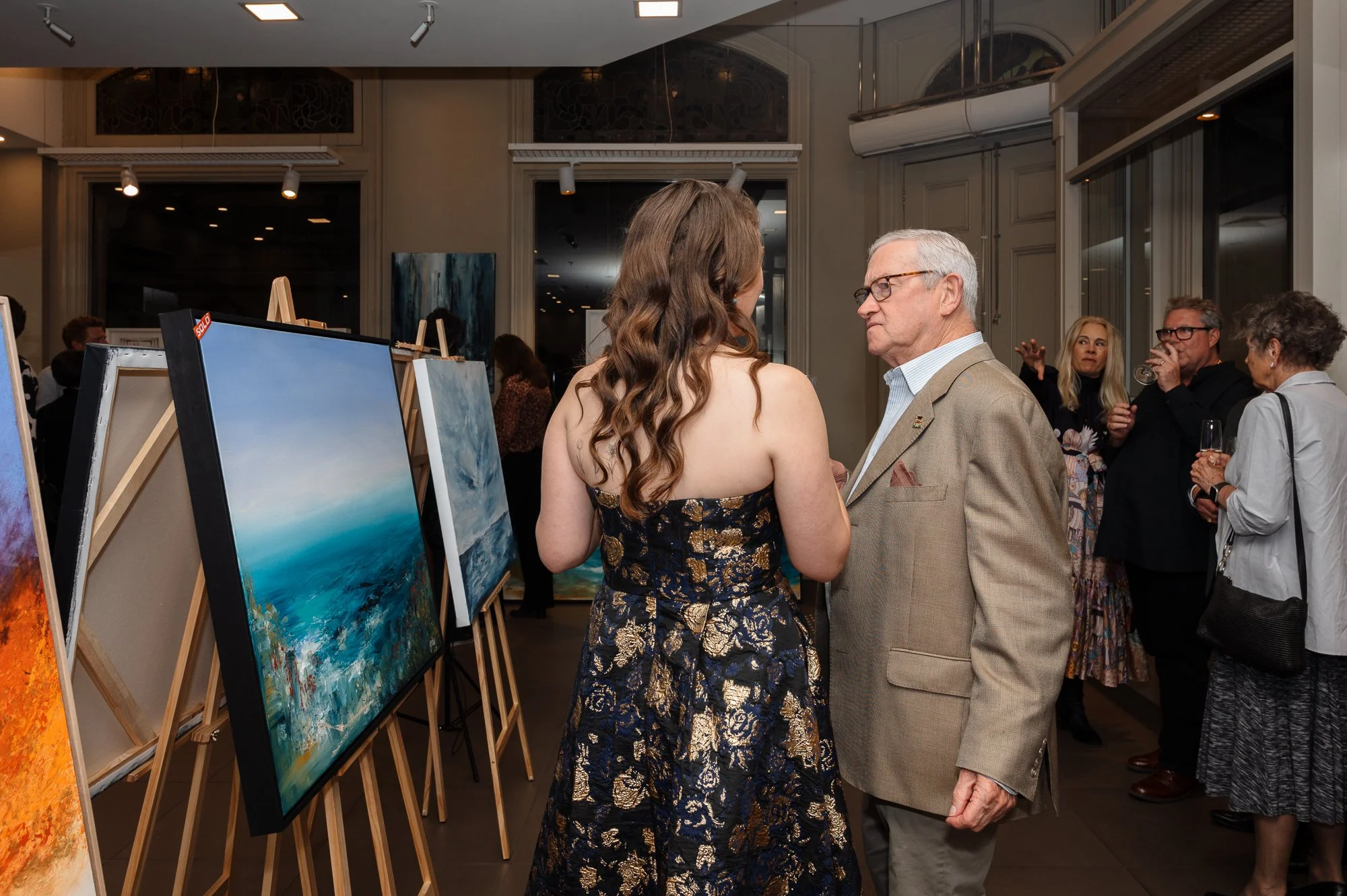 Two people are having a conversation at an art gallery, with paintings displayed on easels around them. The background shows more attendees, some holding drinks, engaging in conversations.