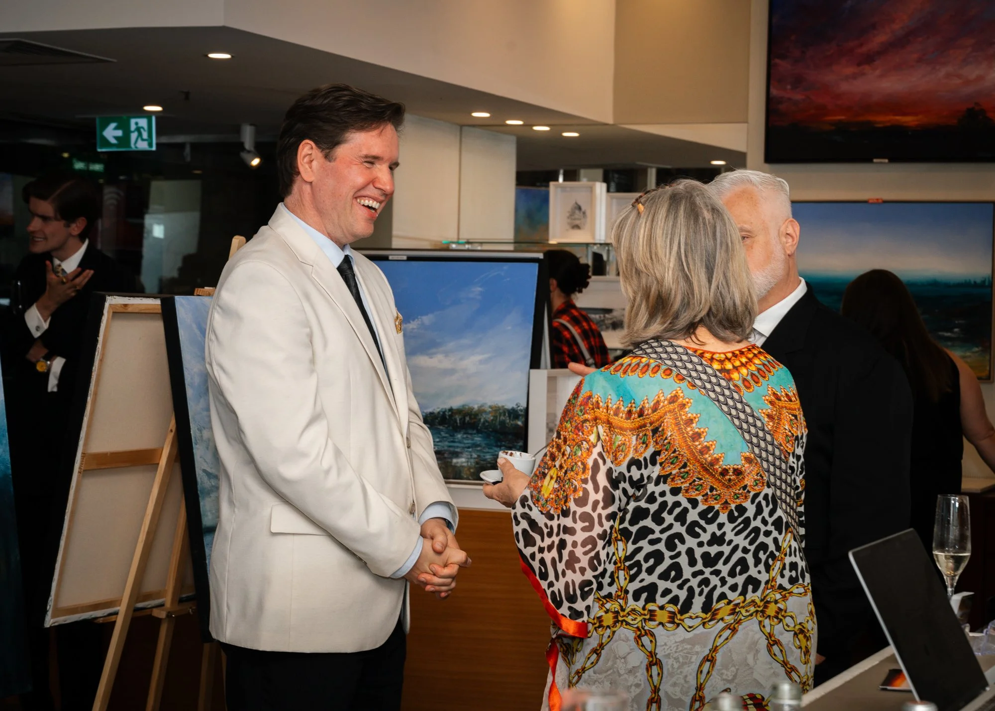 Man in a white suit shaking hands with a woman and talking to a man in a black suit at an art gallery event.