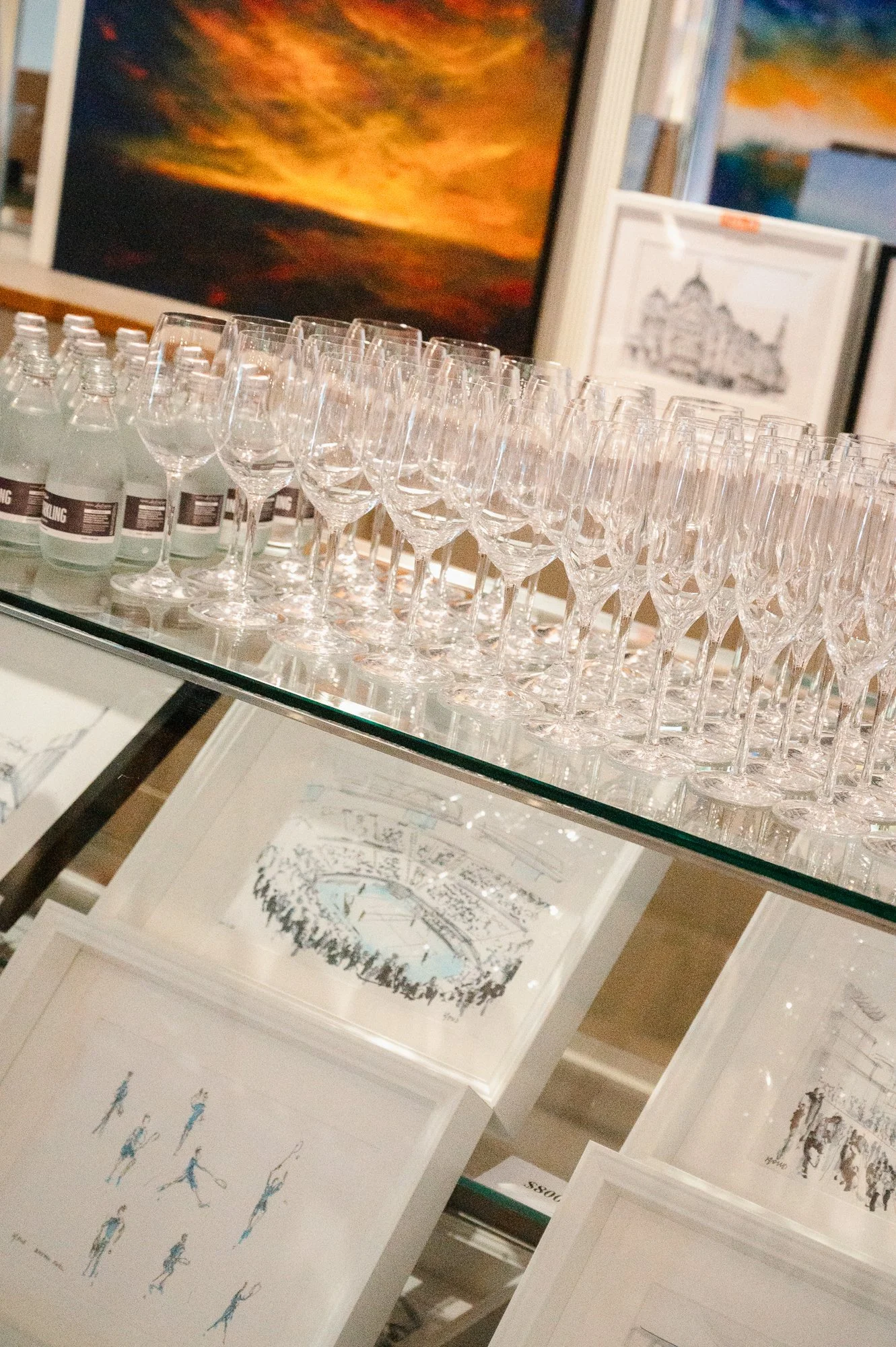 Glassware displayed on a glass shelf with artwork and framed pictures in the background.