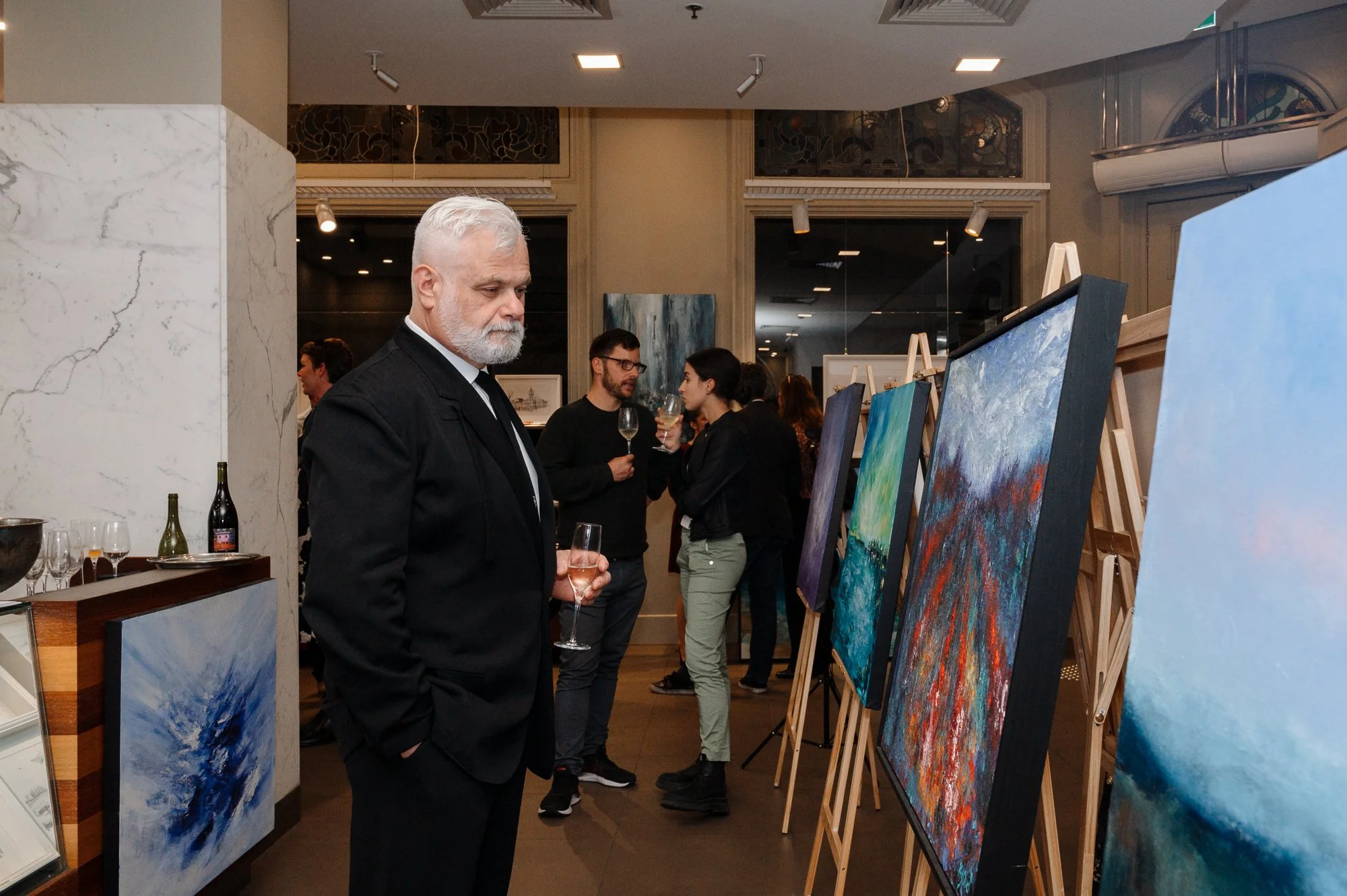 An art gallery opening with people viewing abstract paintings on display. A man with white hair and beard in a black suit holds a glass of champagne, observing the artwork. Other attendees are mingling and holding drinks in the background.